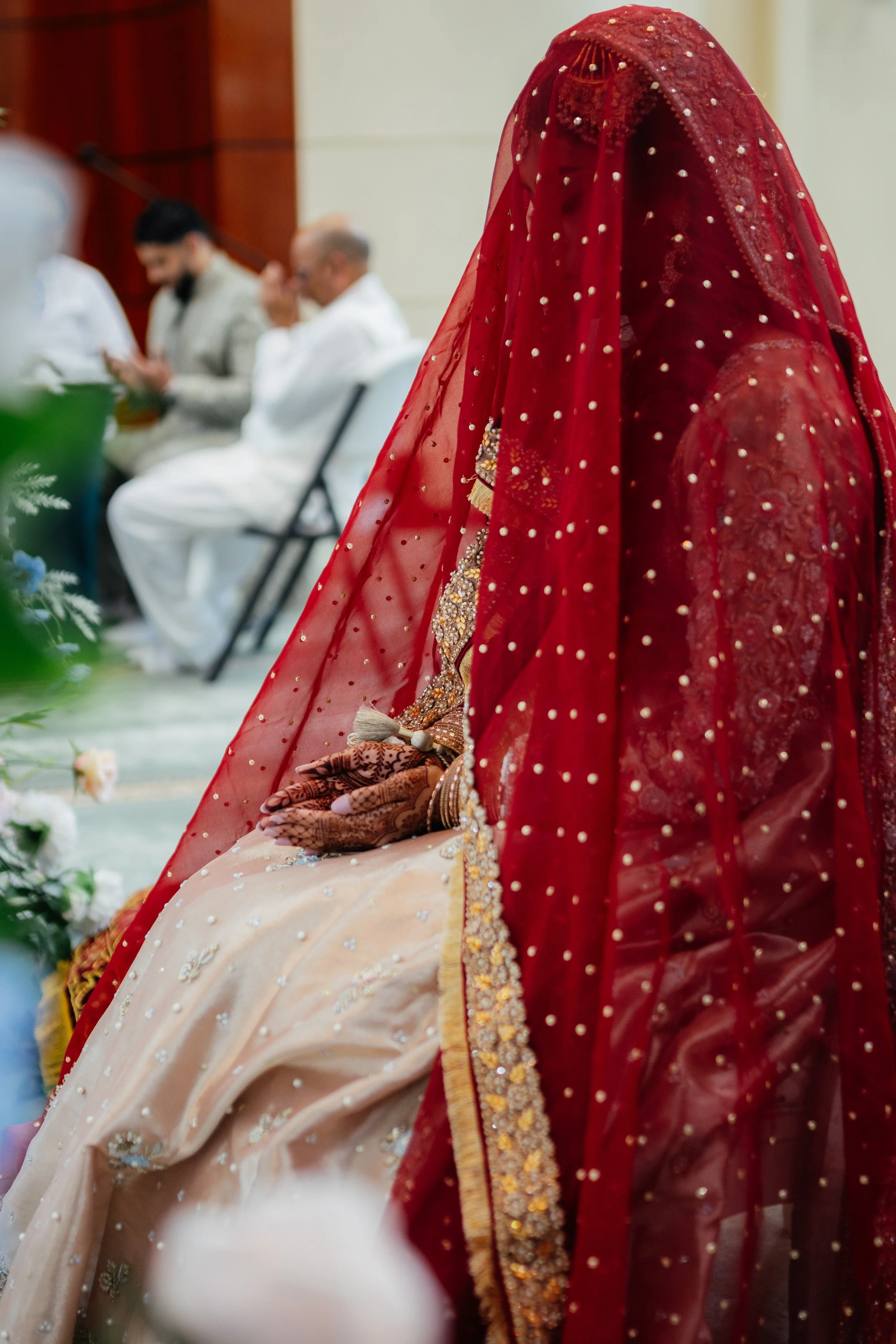 A woman dressed in traditional red and beige Indian attire with intricate embroidery, sitting with hands clasped. She is wearing a red dupatta with embroidered details, covering her head and face. In the background, three men are seated, dressed in w