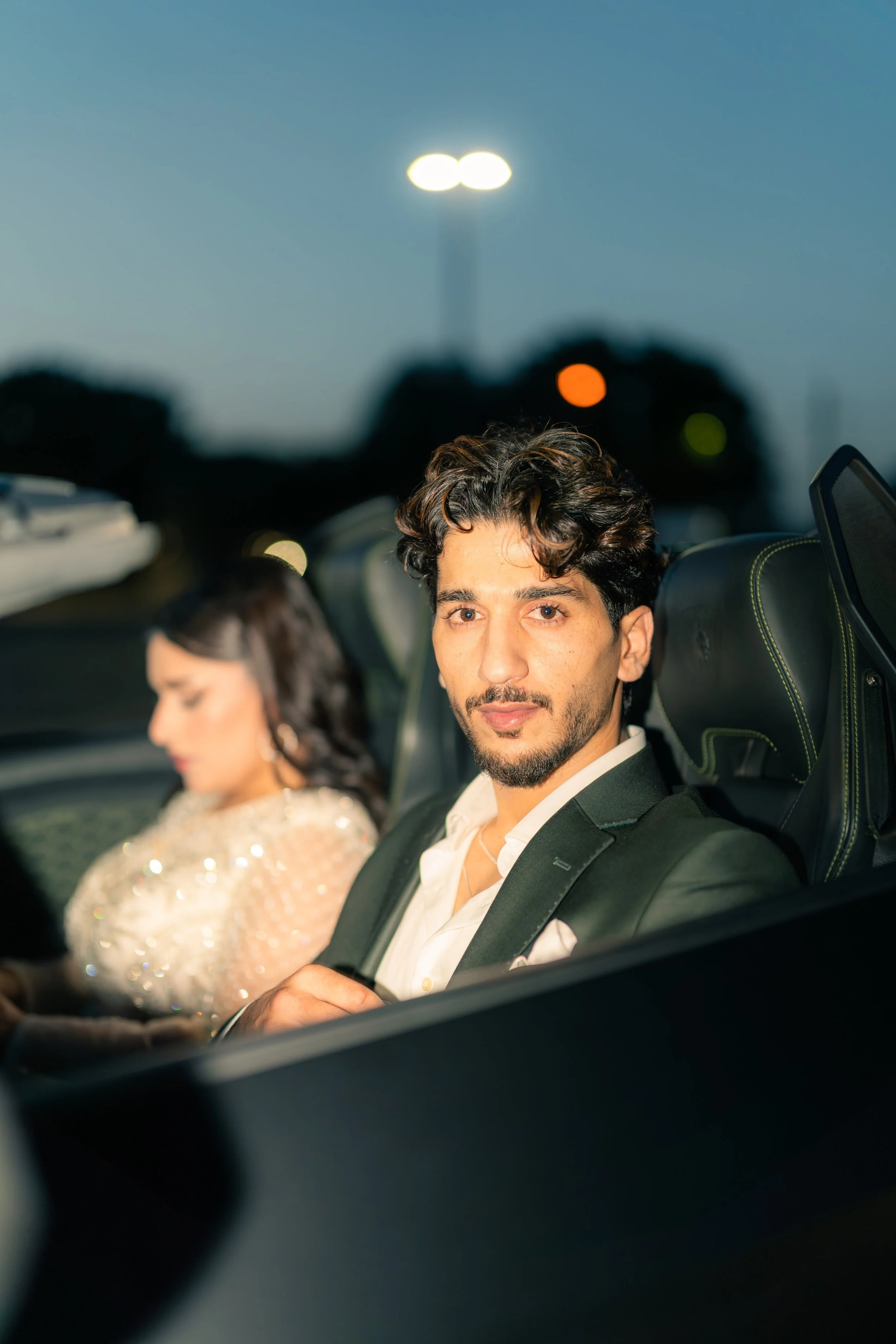 A man in a black suit and white shirt sitting in the backseat of a luxury car with a woman in a sparkling dress sitting next to him, blurred in the background, during dusk.
