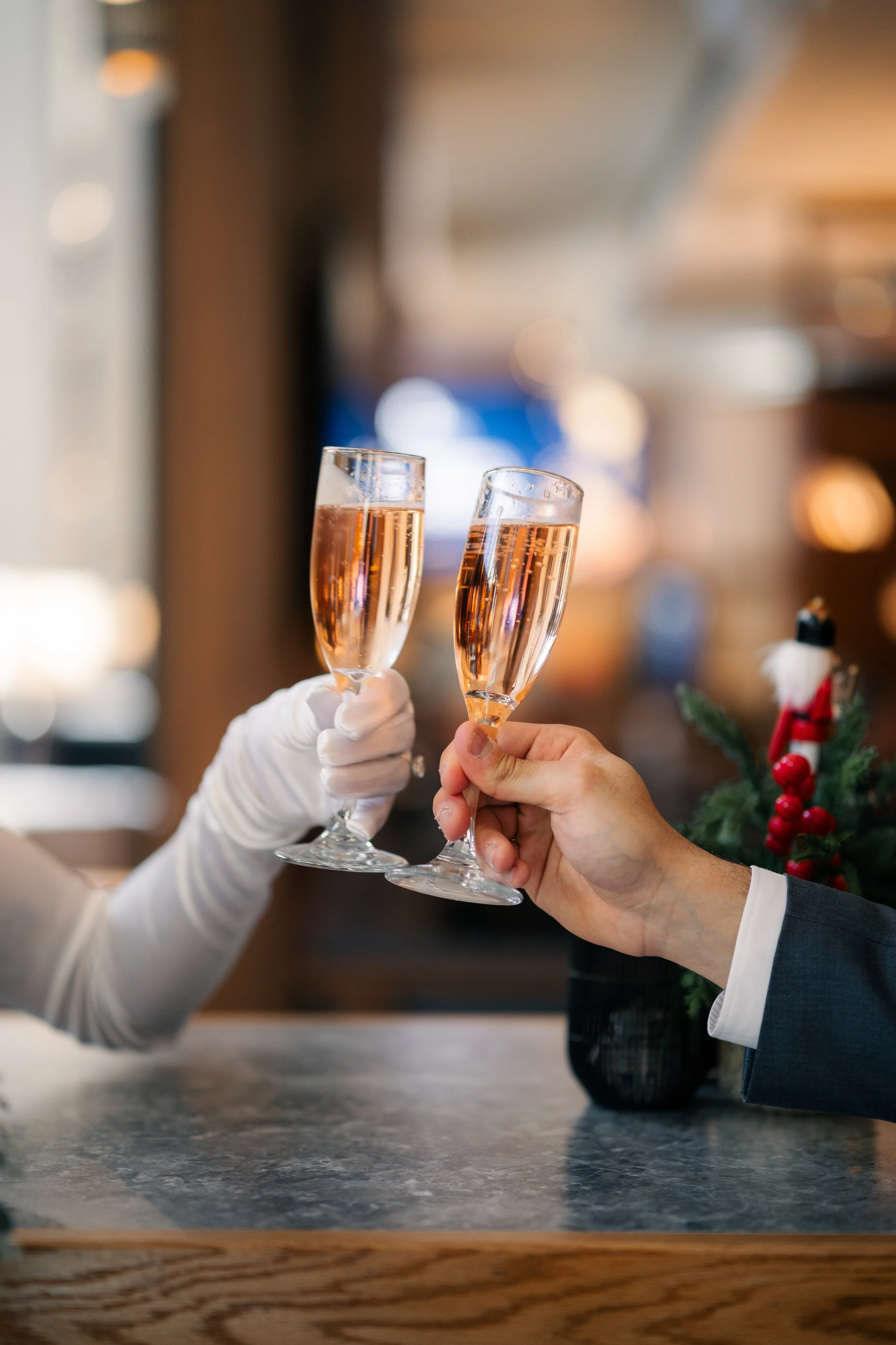 Two people clinking glasses of rosé champagne, one person wearing a white glove, celebrating at a festive event, with holiday decorations nearby.
