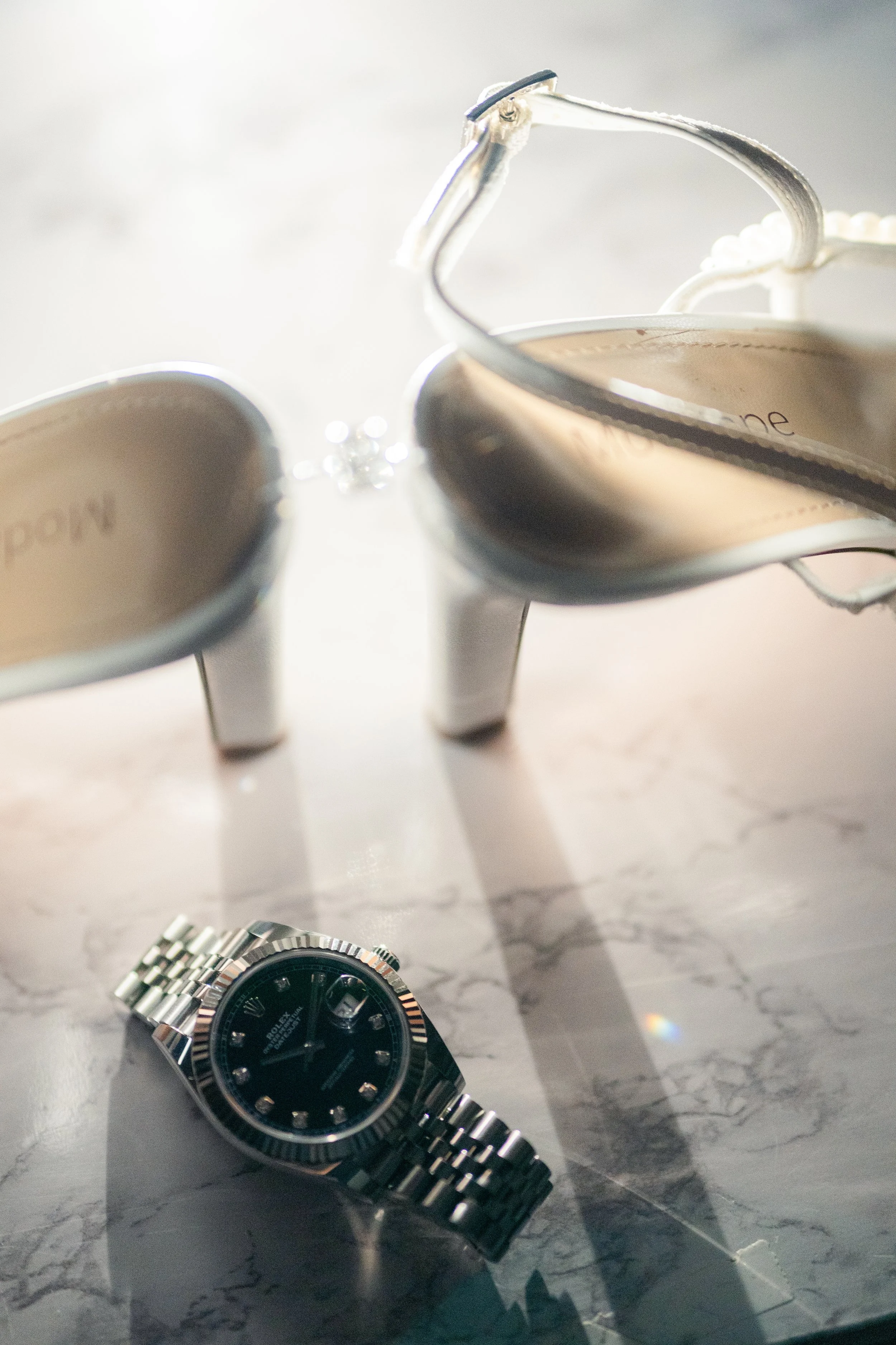 A pair of silver high-heeled shoes, a wristwatch with a black face and silver band, and a pearl necklace on a marble surface.