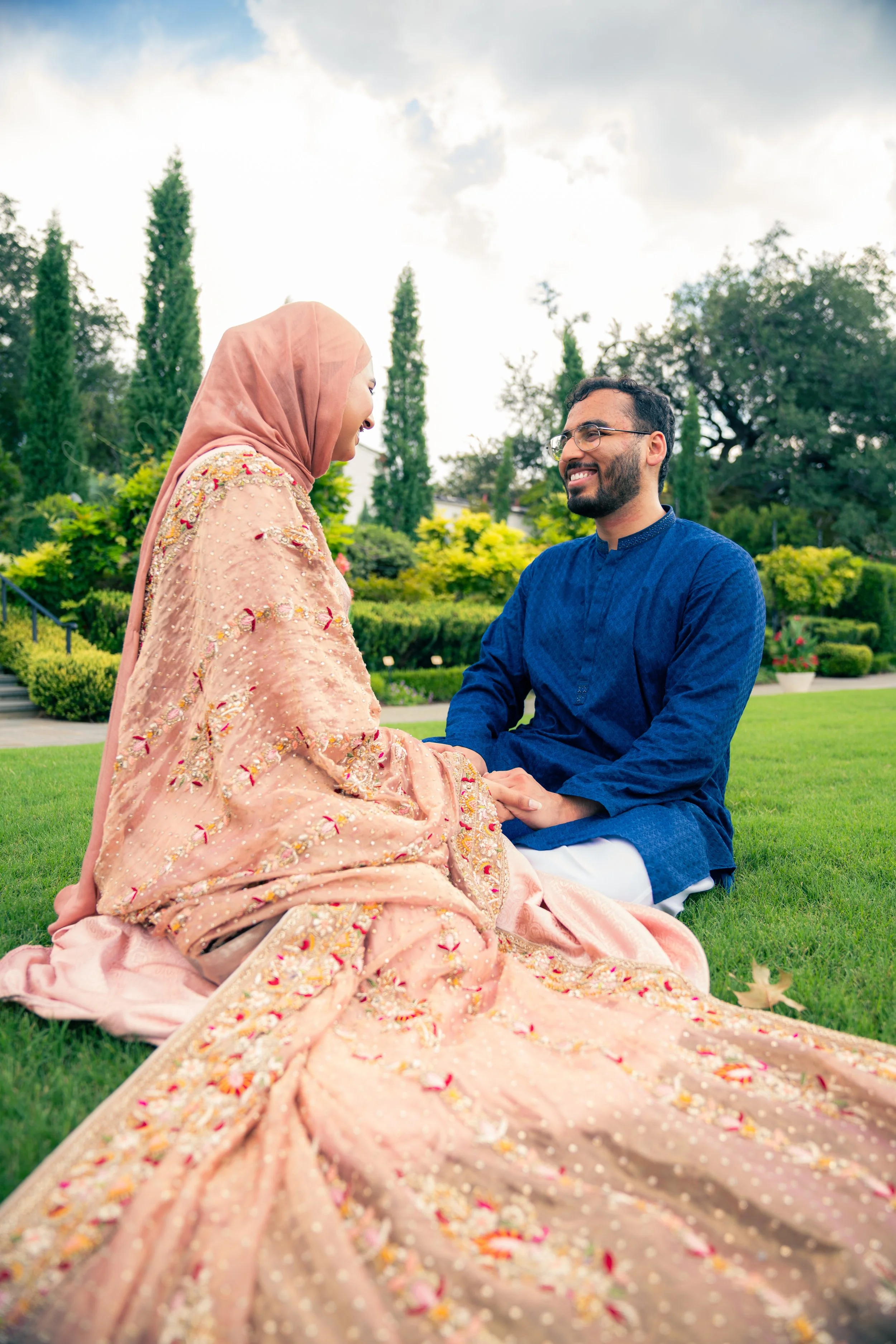 A couple dressed in traditional South Asian attire sitting on the grass in a park, holding hands and smiling at each other.