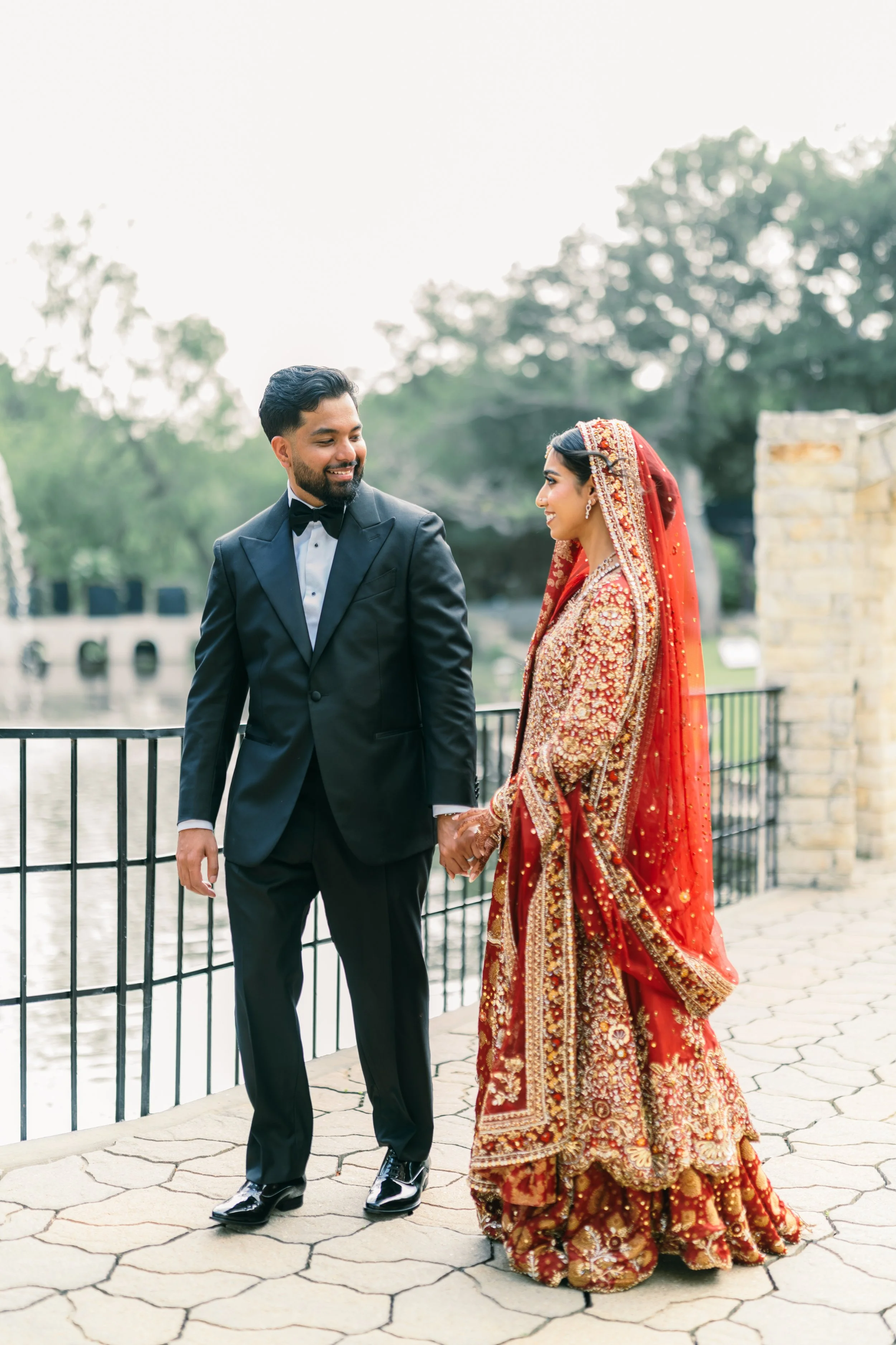 A groom in a black tuxedo holding hands with a bride in a red and gold traditional South Asian wedding attire, both smiling and walking outdoors near a railing by a body of water with trees in the background.