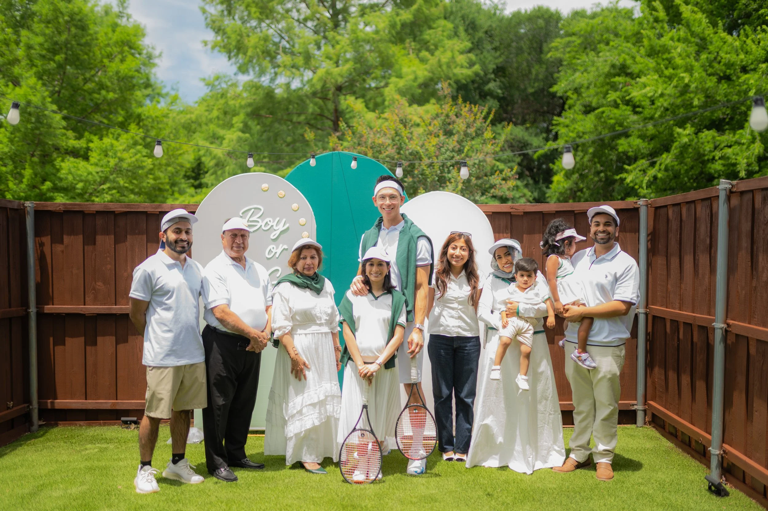 A group of diverse people at a backyard tennis-themed gathering, some holding tennis rackets, with a sign reading 'Boy or Girl' behind them, plants, trees, and string lights in the background.