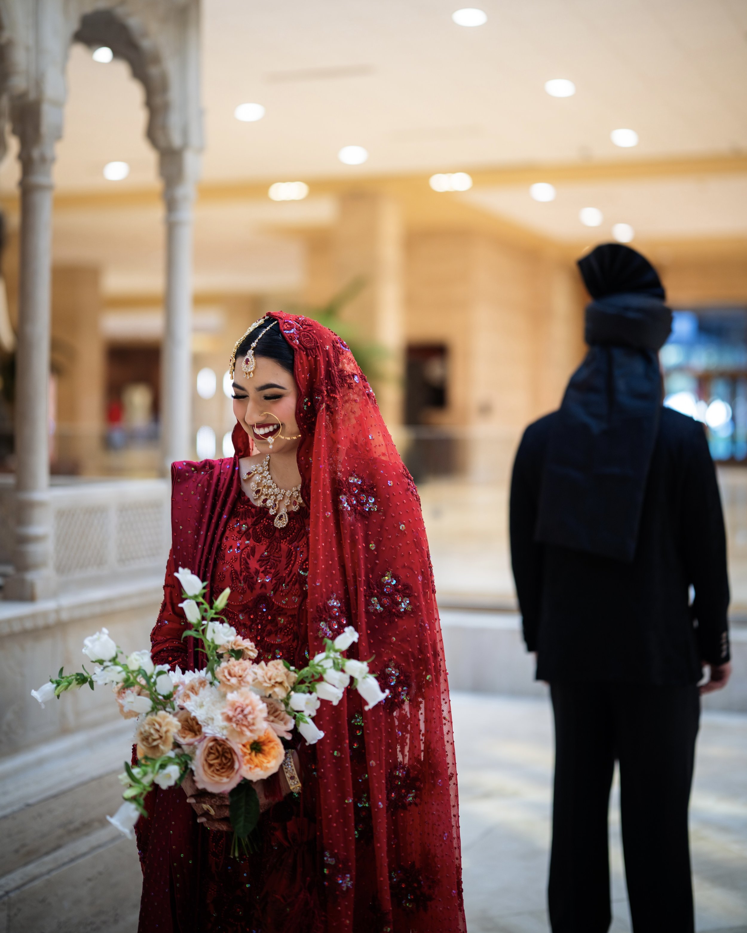 A woman in a red traditional dress and veil, smiling and holding a bouquet of flowers, standing indoors with a man in dark clothing and a turban in the background.