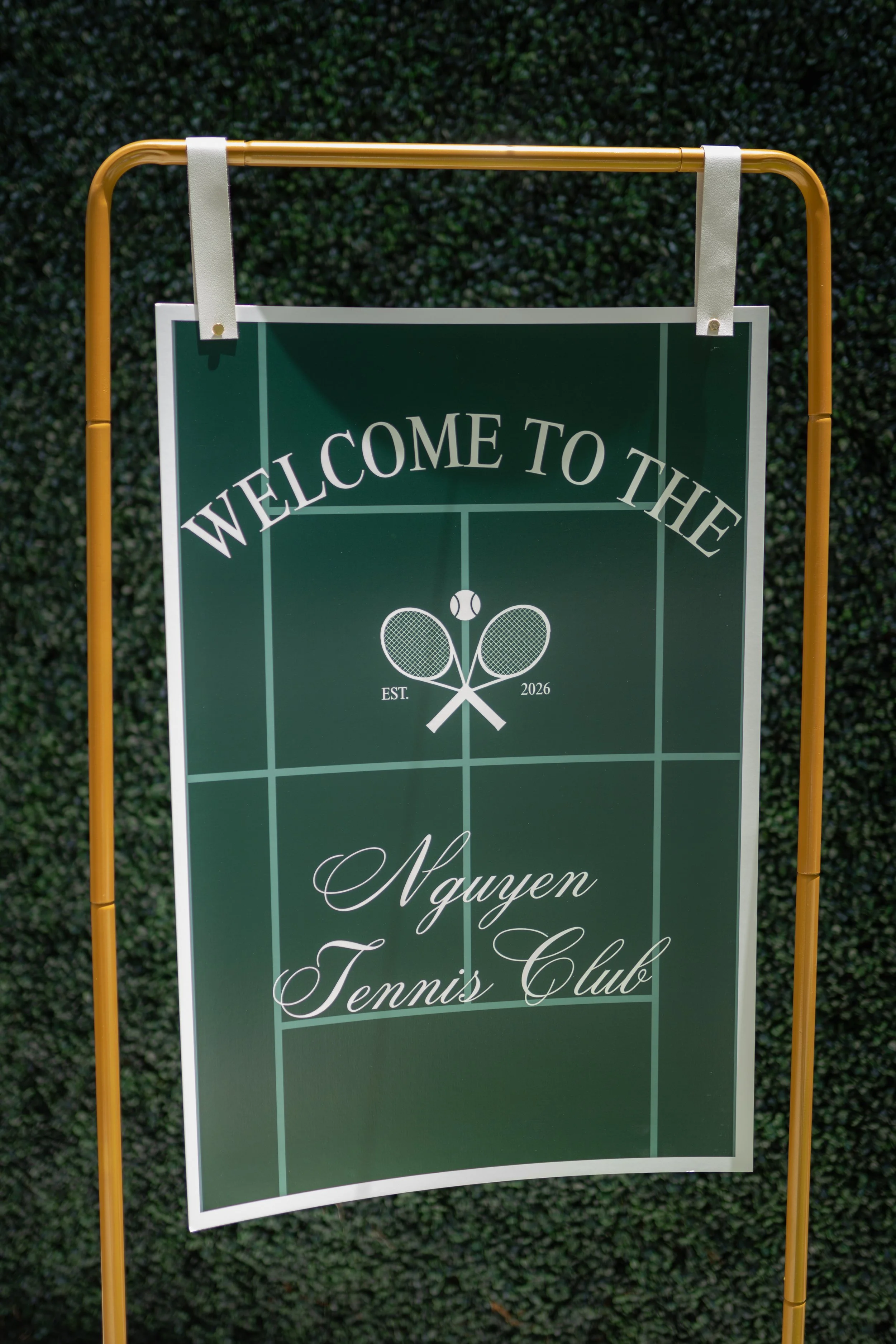 A green sign with white text that reads "Welcome to the" and includes a tennis theme with crossed tennis rackets and a tennis ball, indicating a tennis club established in 2026, with the name "Nguyen Tennis Club".