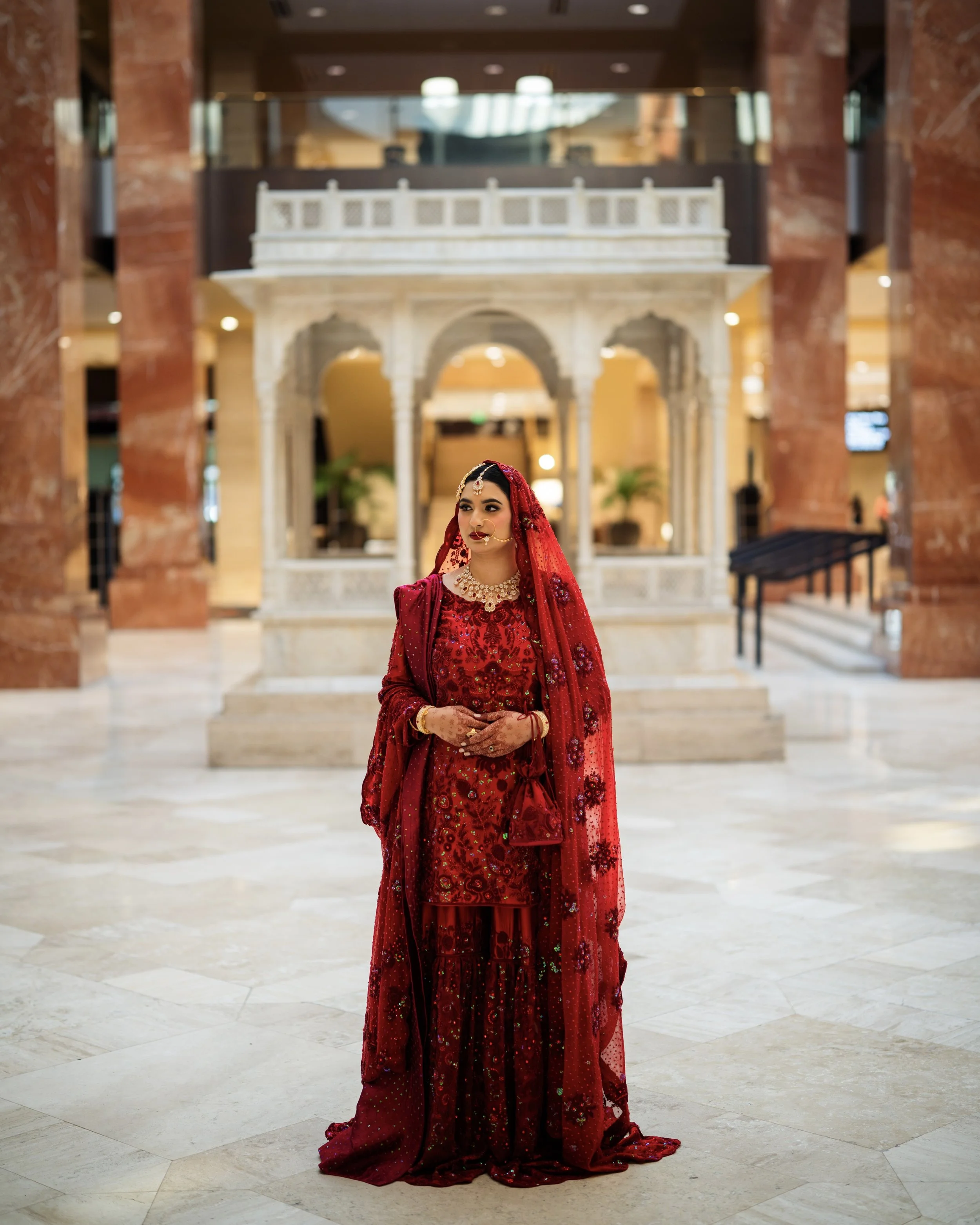 A woman dressed in traditional red bridal attire standing in a spacious, elegant hall with marble flooring and a decorative white marble structure in the background.