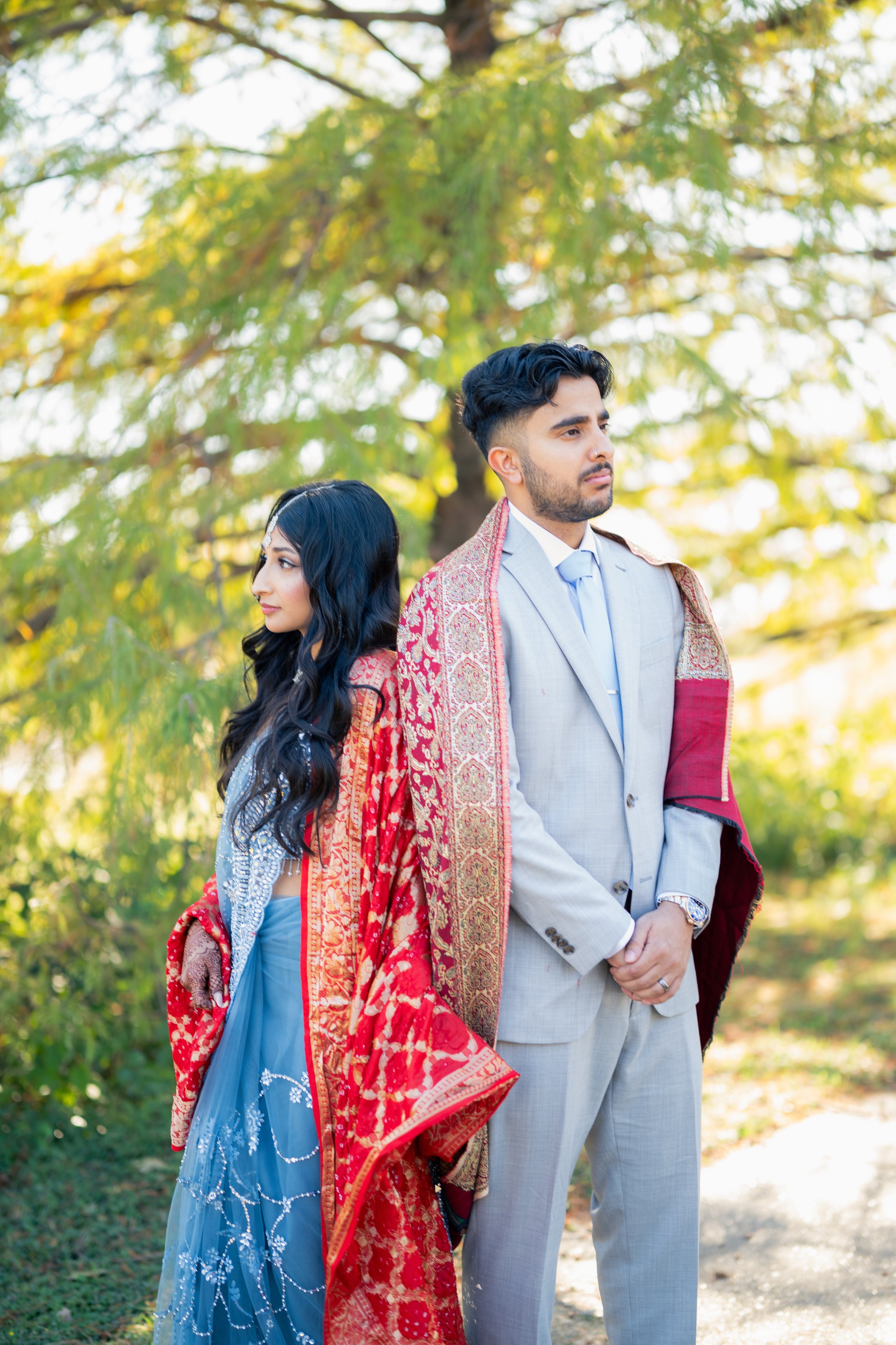A man and woman dressed in formal and traditional attire standing outdoors near green trees, with a woman in a colorful traditional outfit and a man in a light-colored suit, both looking away from each other.