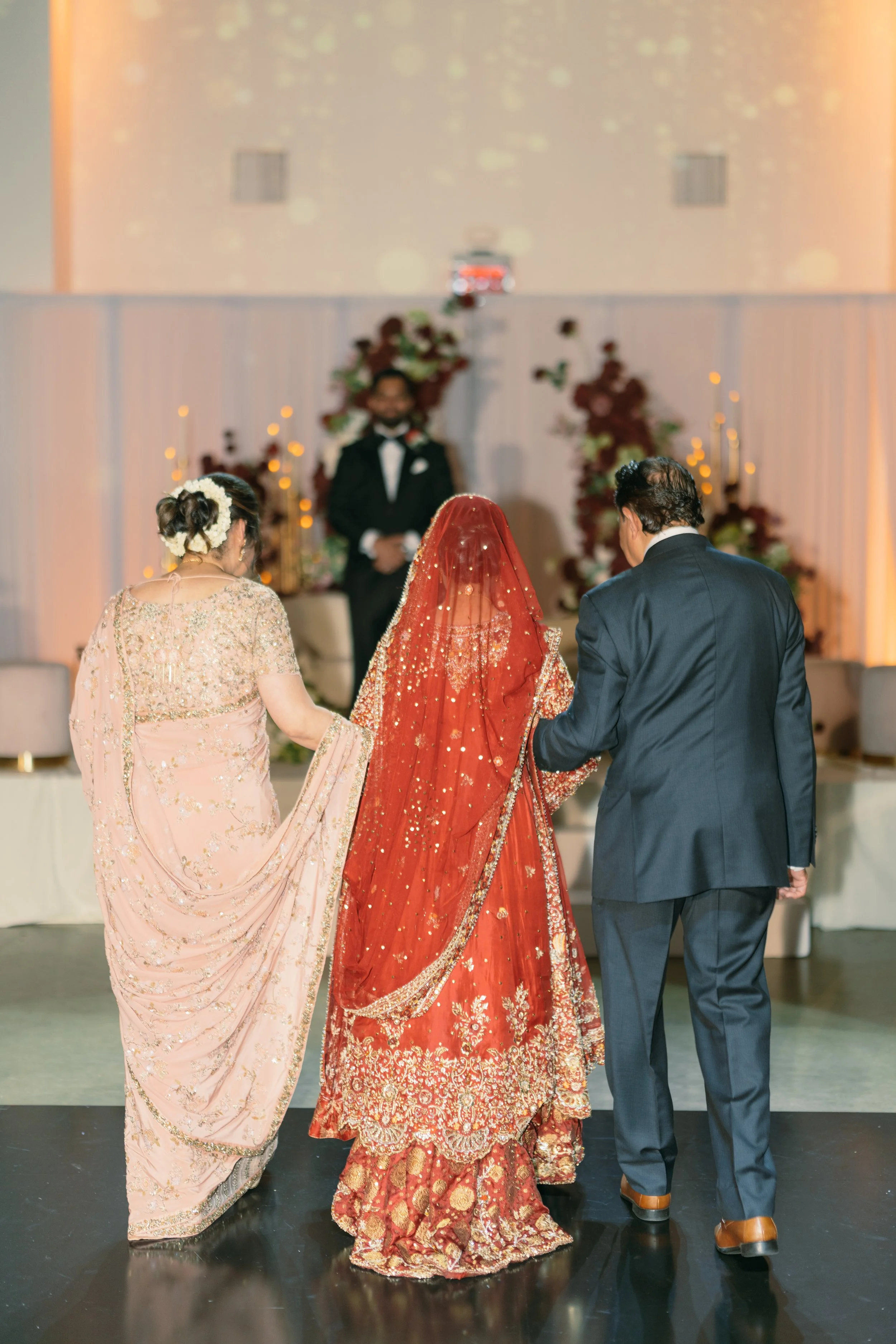 A bride in a red and gold traditional wedding dress walking down the aisle with her parents at a wedding ceremony.