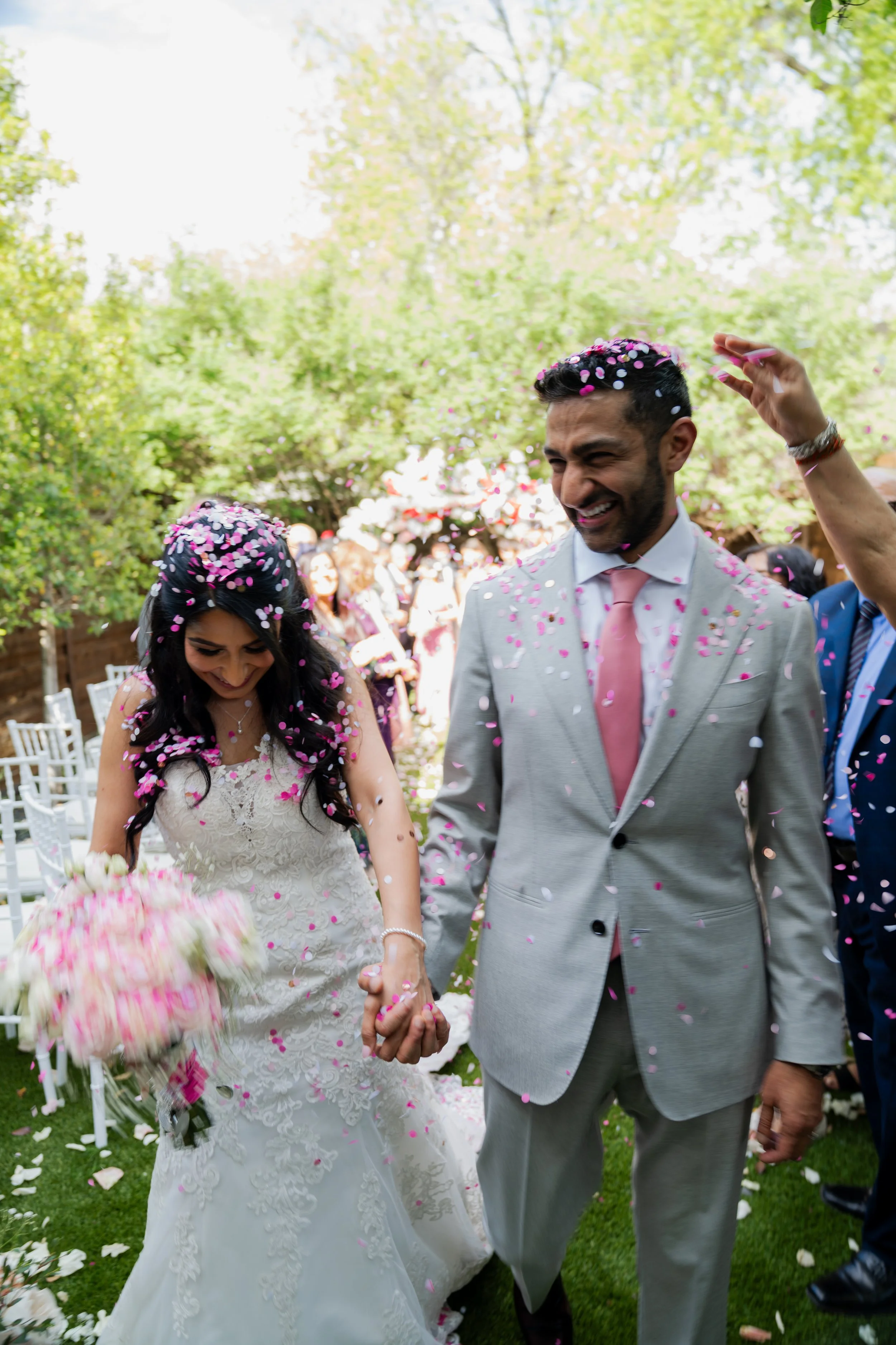 A newlywed couple holding hands and smiling during their outdoor wedding ceremony. The bride is wearing a white lace wedding dress and holding a bouquet, while the groom is in a light grey suit with a pink tie. Pink and white confetti is falling arou