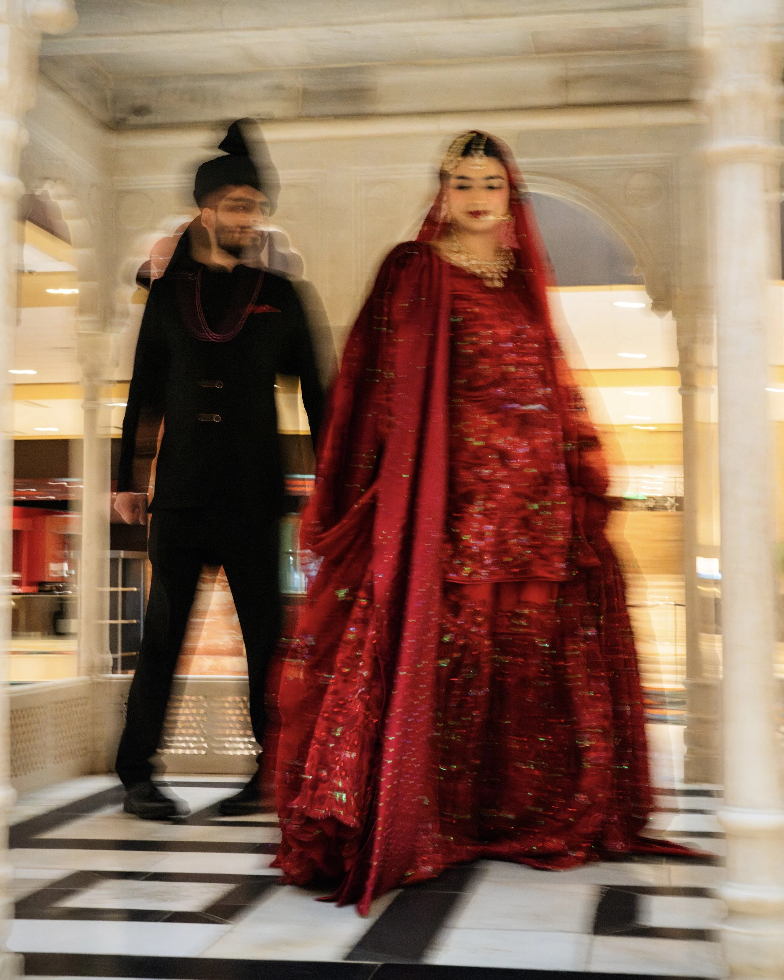 A woman in a vibrant red traditional outfit with gold embroidery and jewelry, walking with a man in a black suit and turban inside an elegant marble-clad building.