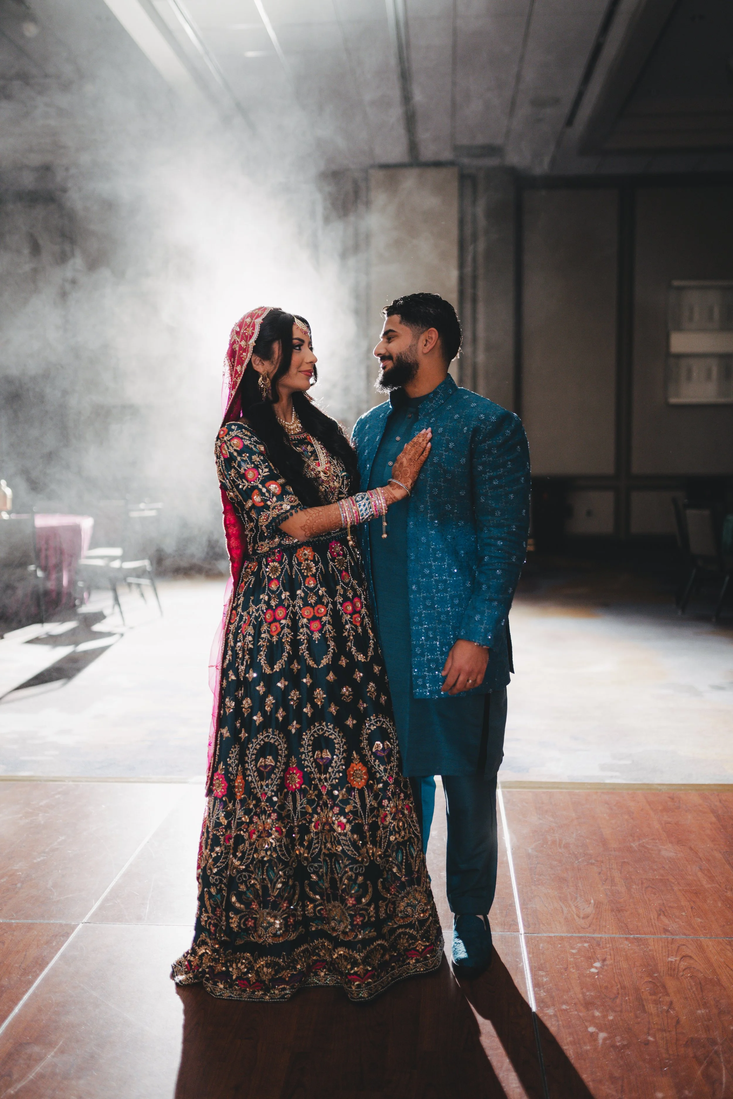 A couple dressed in traditional South Asian wedding attire standing face to face, gazing into each other's eyes in a decorated indoor venue with natural light.