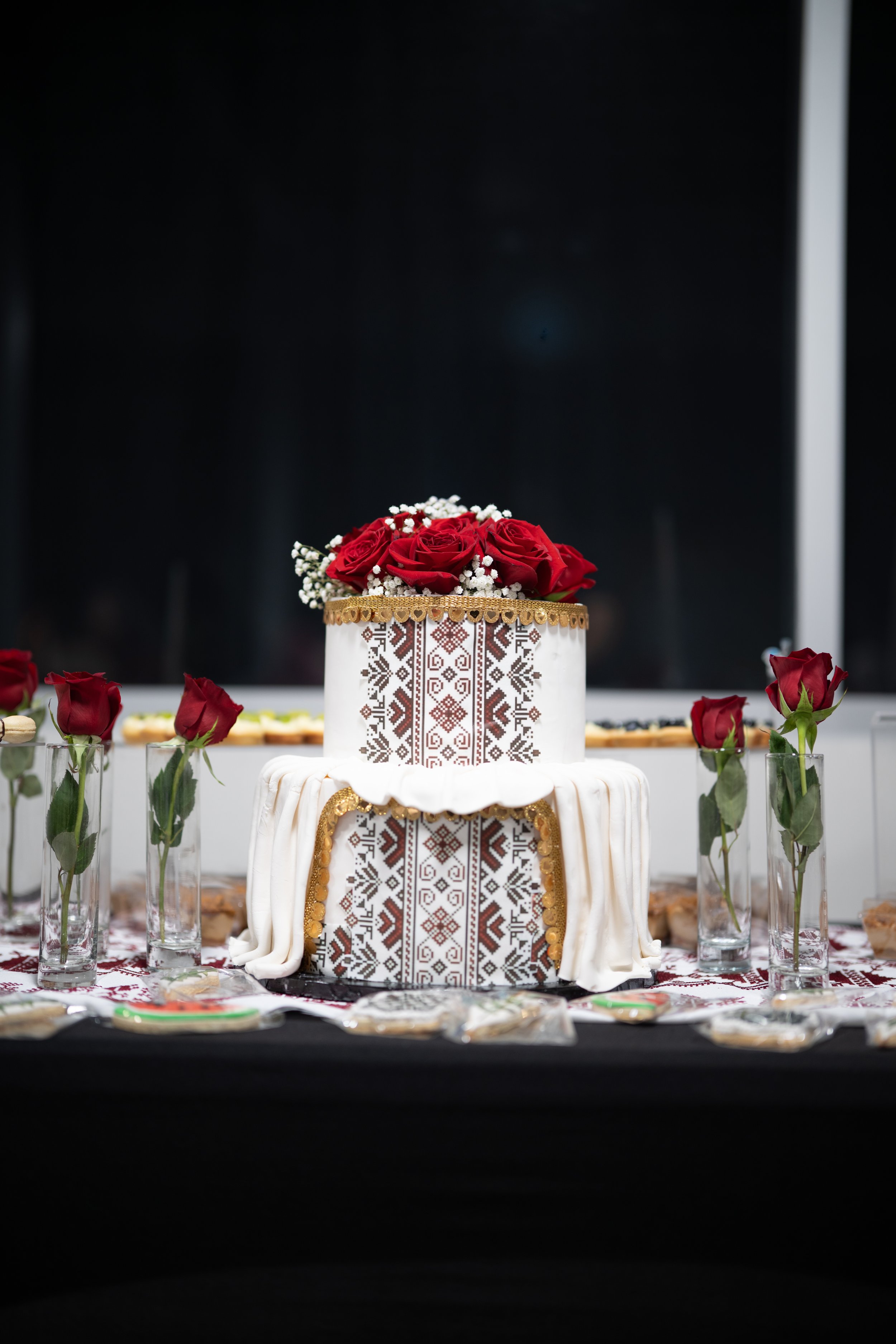 Two-tiered wedding cake decorated with traditional embroidery patterns, topped with red roses and small white flowers, surrounded by red roses in glass vases