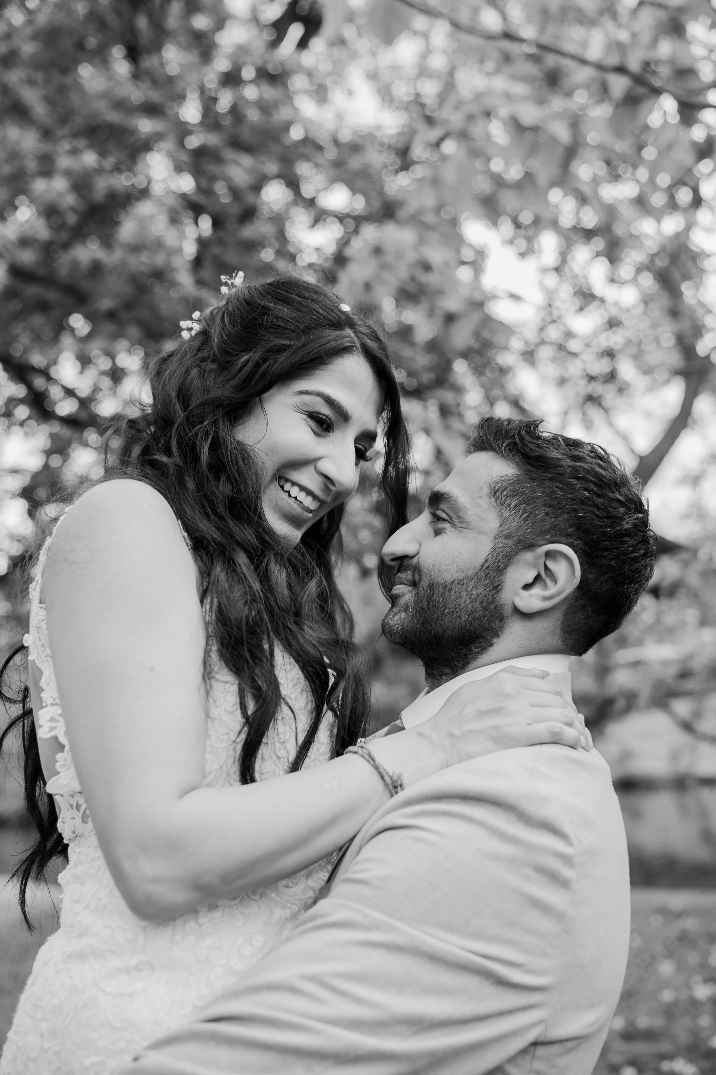 A black and white photo of a smiling woman and a man looking at each other closely outdoors, with trees and blurred background.