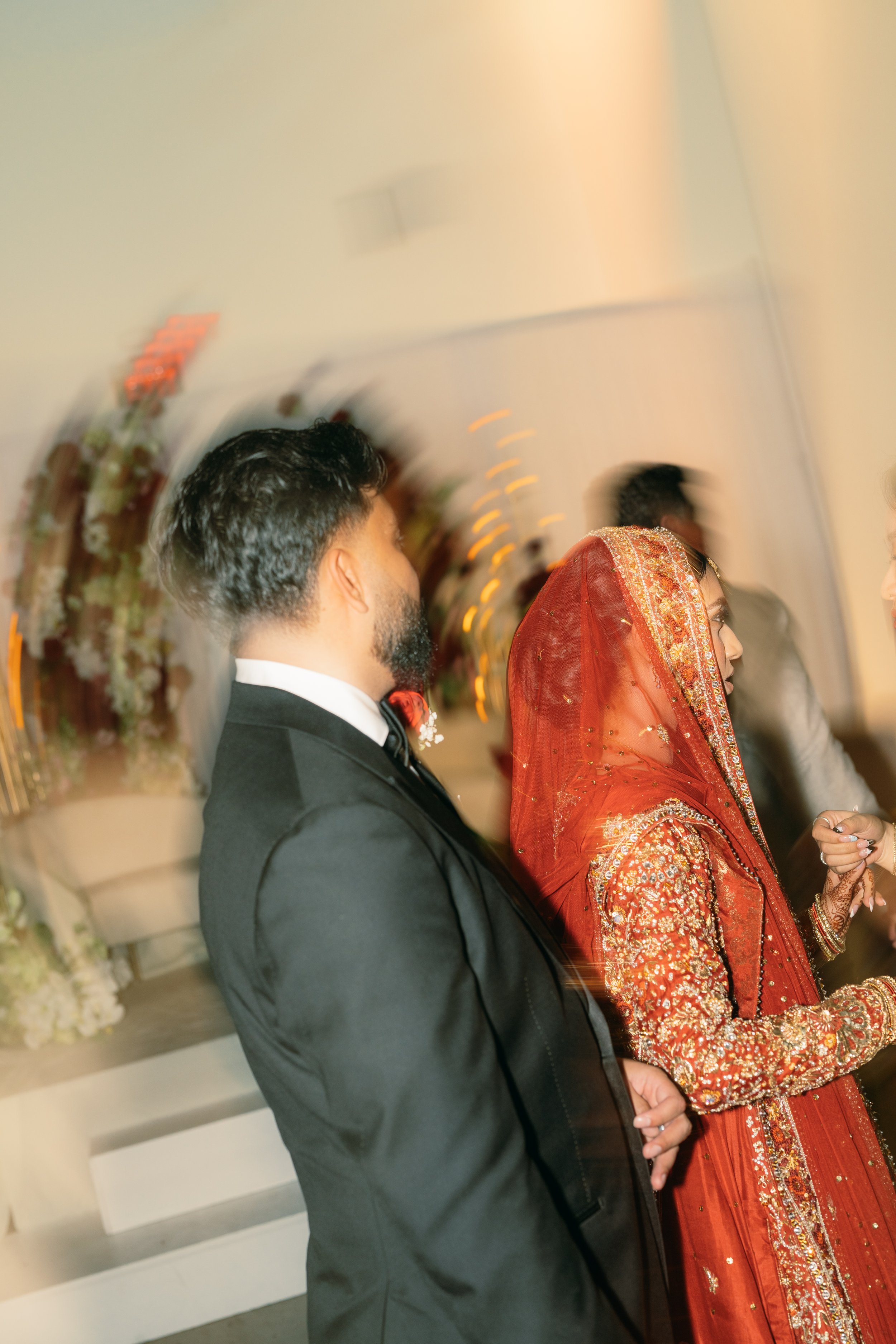A couple at a wedding, with the man in a black tuxedo and the woman dressed in a traditional red and gold Indian saree, standing indoors under soft lighting, with floral decorations in the background.