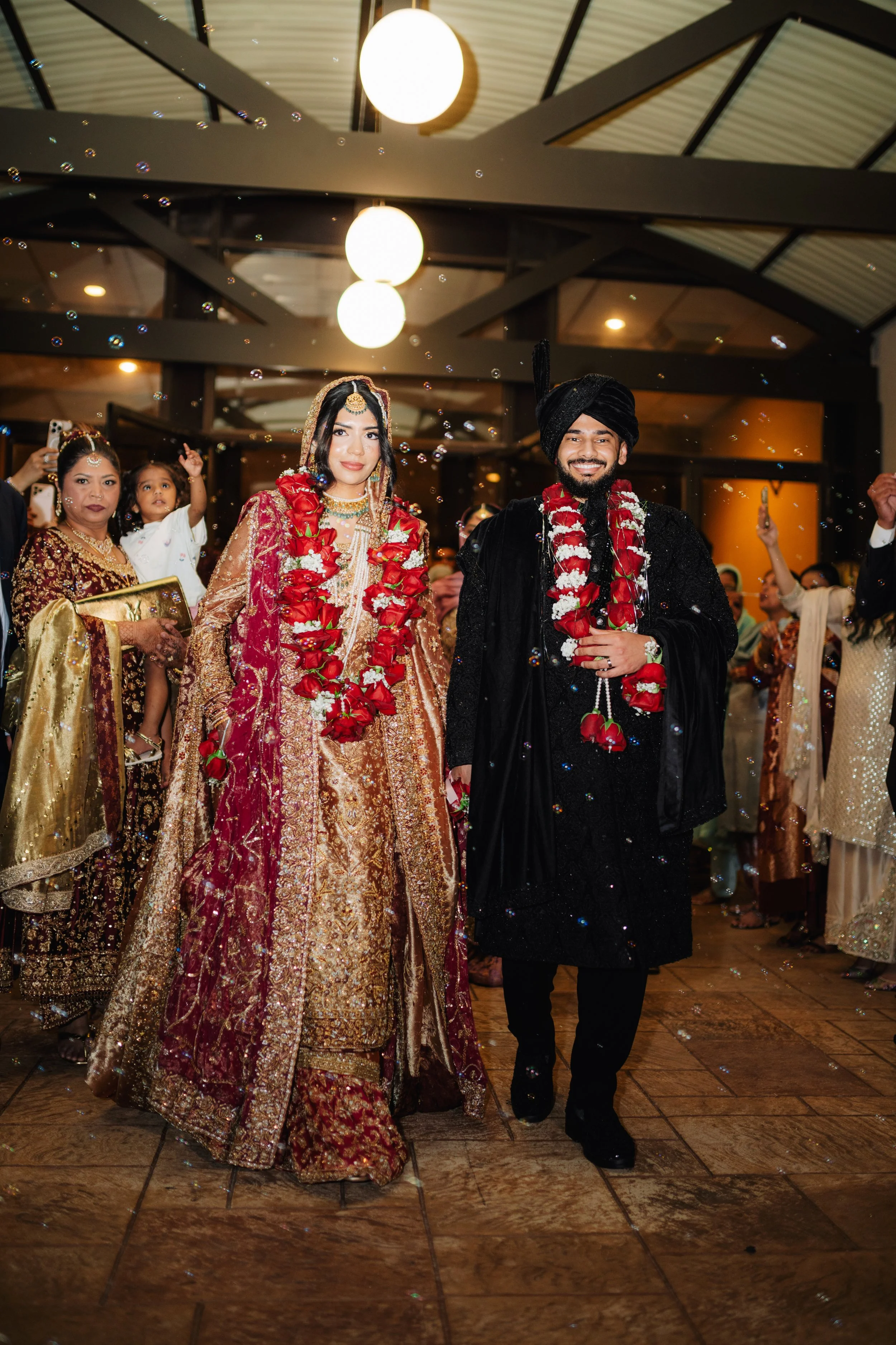 A South Asian wedding reception with the bride and groom dressed in traditional attire, walking together and smiling. The bride wears a richly embroidered maroon and gold saree with jewelry, and the groom wears a black sherwani with a matching turban