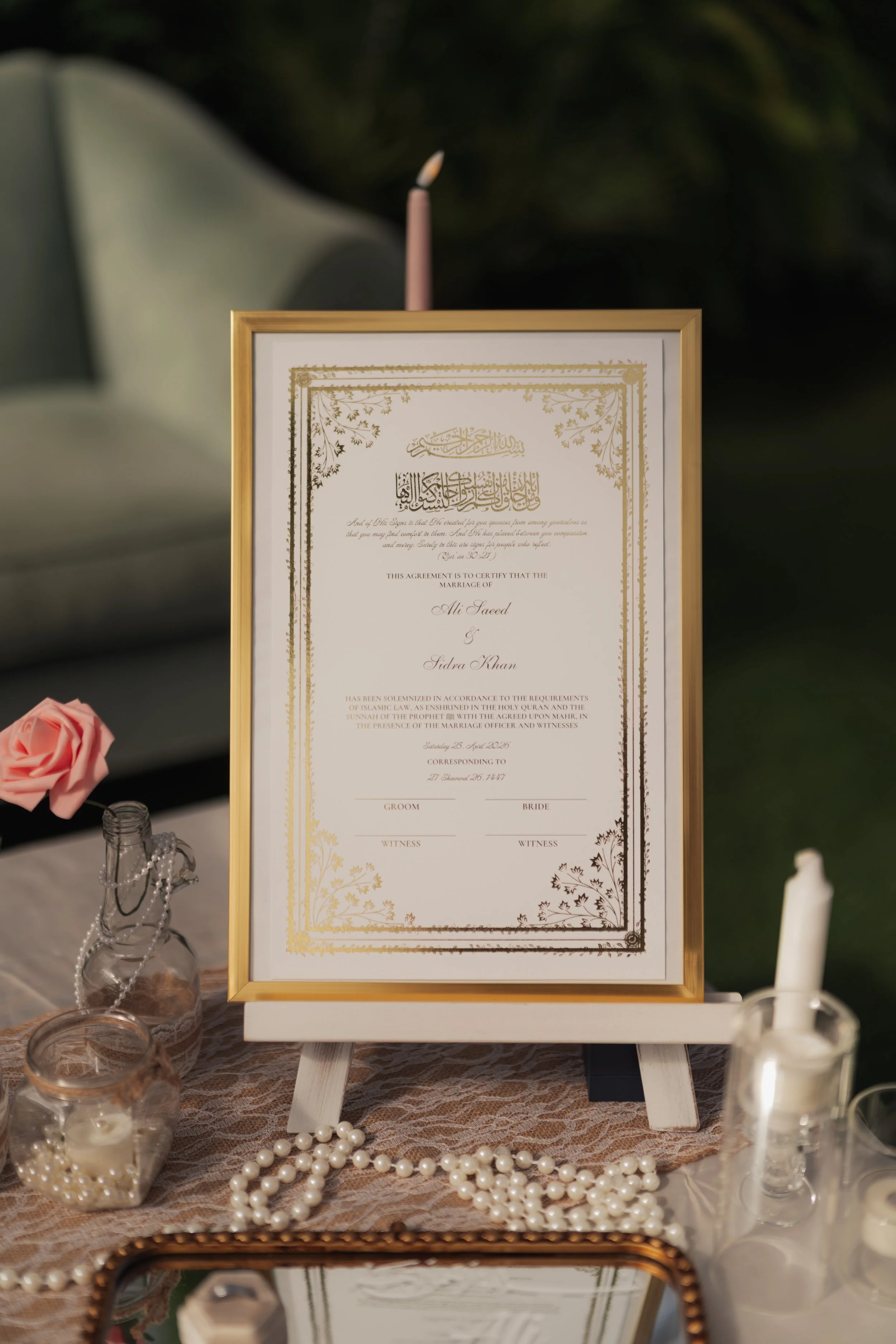 A framed marriage certificate displayed on a table with decorative candles, pearl necklaces, and pink roses, set against a dark background.