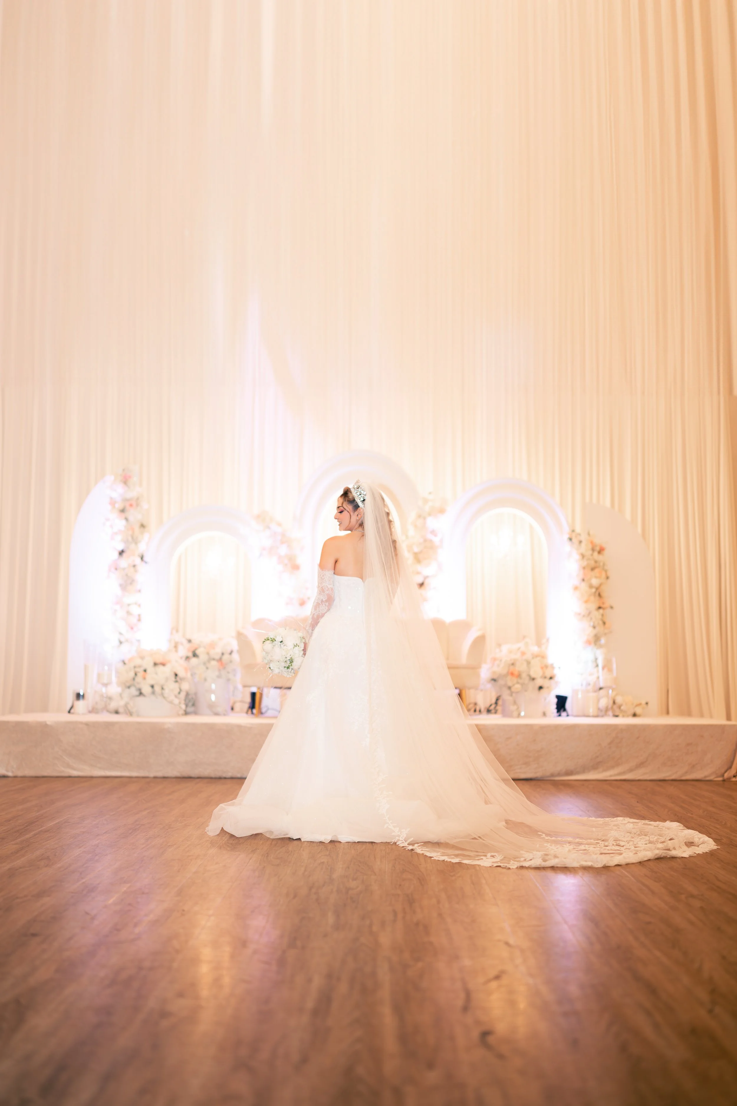 A bride in a white wedding dress holding a bouquet stands in a decorated wedding hall with floral arrangements and soft lighting.