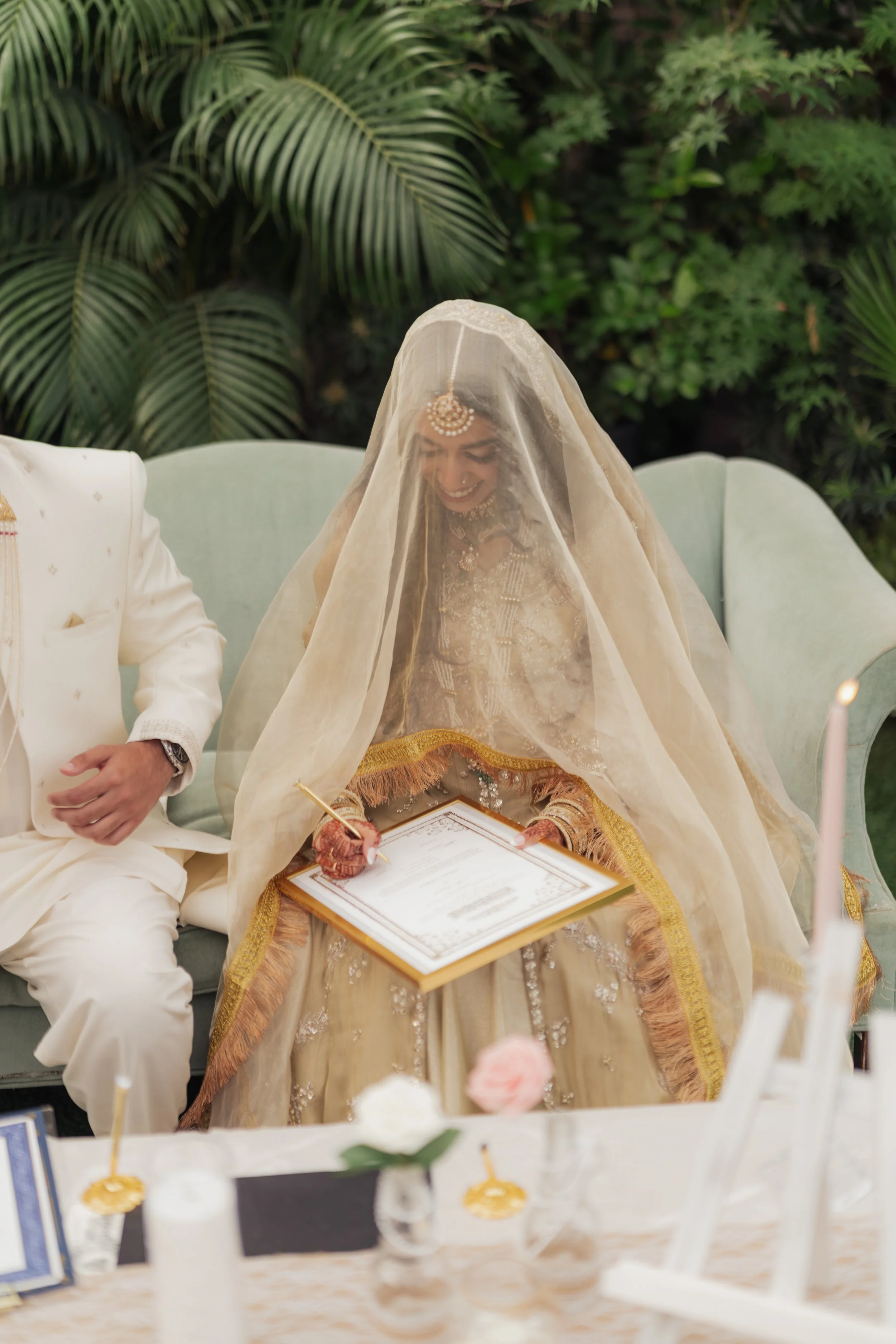 A woman dressed in traditional Indian wedding attire sitting at a reception table with a veil over her face, holding a certificate and smiling. She is seated next to a man in a cream-colored suit, and the setting has lush green plants in the backgrou