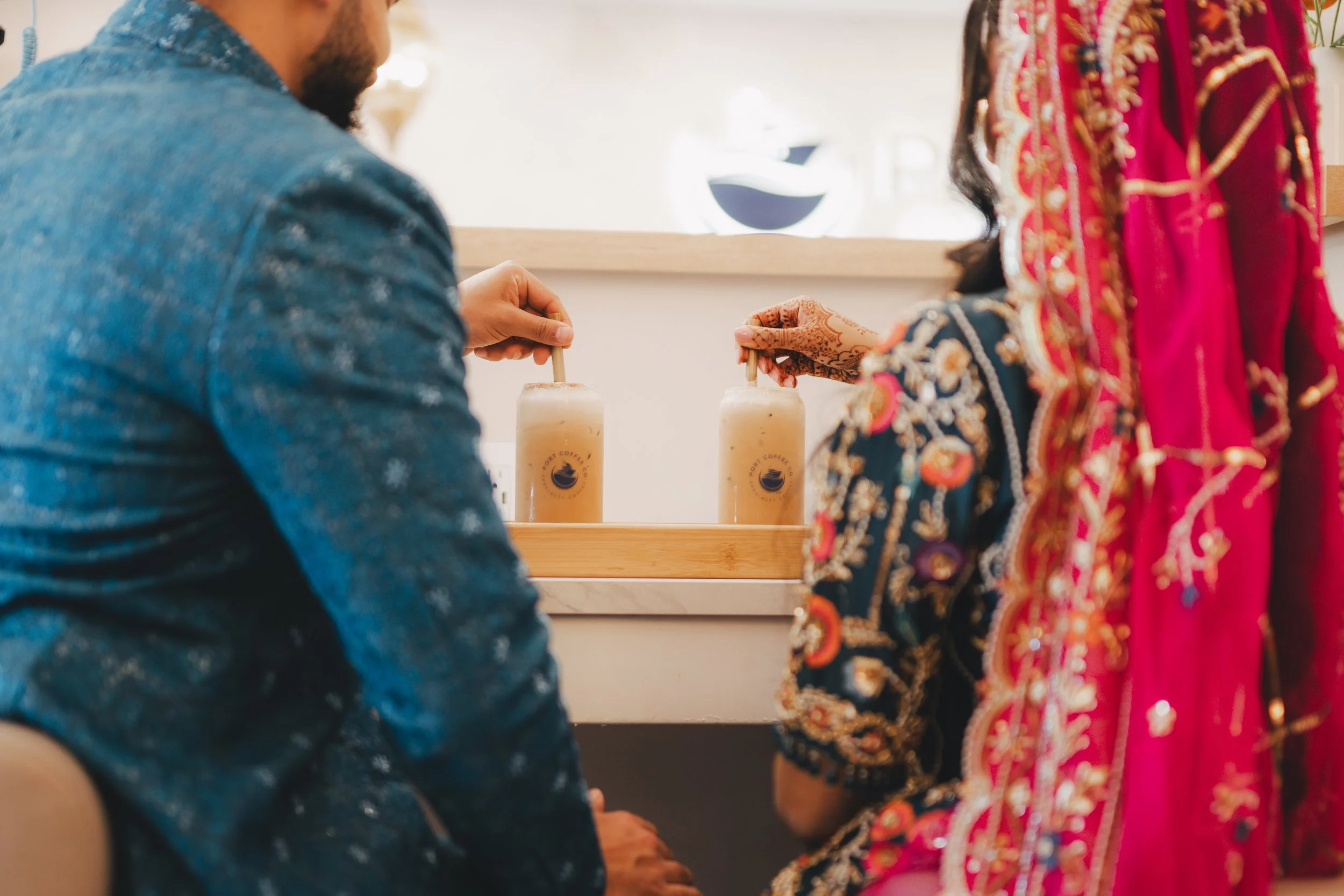 A man in a blue denim shirt and a woman in a colorful embroidered traditional outfit are holding iced coffee drinks with straws inside a cafe.