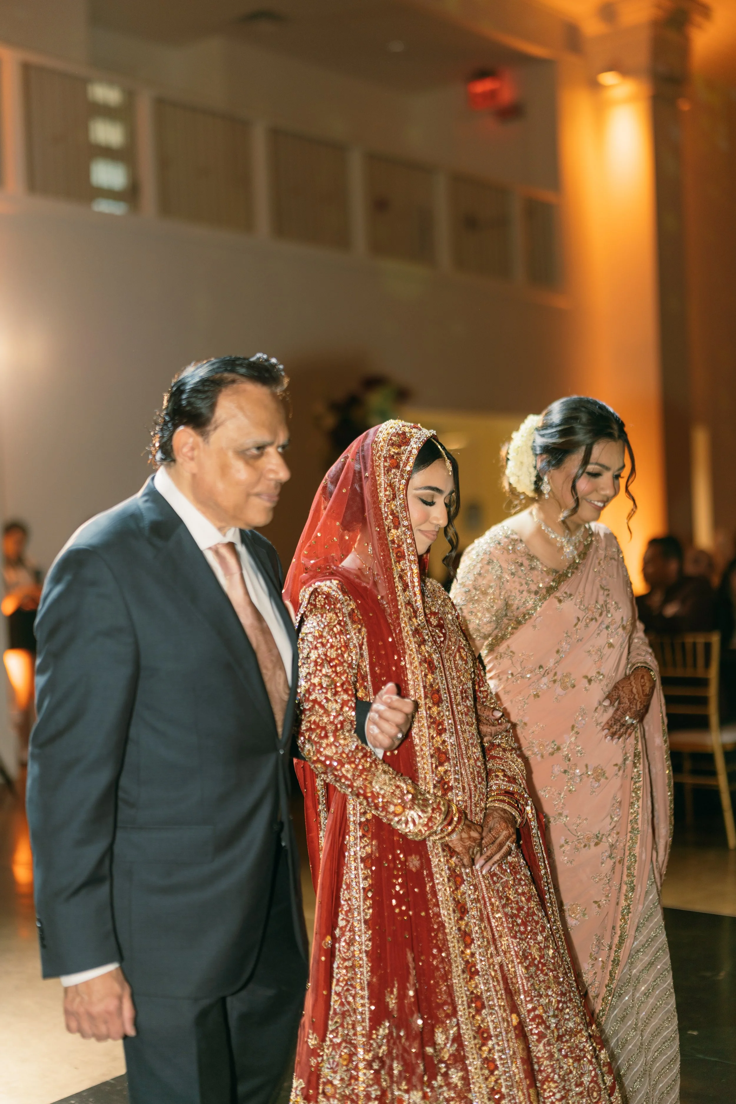 Three people in formal attire standing side by side at an indoor event. The man on the left is wearing a dark suit with a white shirt and silk tie. The woman in the middle is dressed in traditional South Asian wedding attire, with a heavily embroider