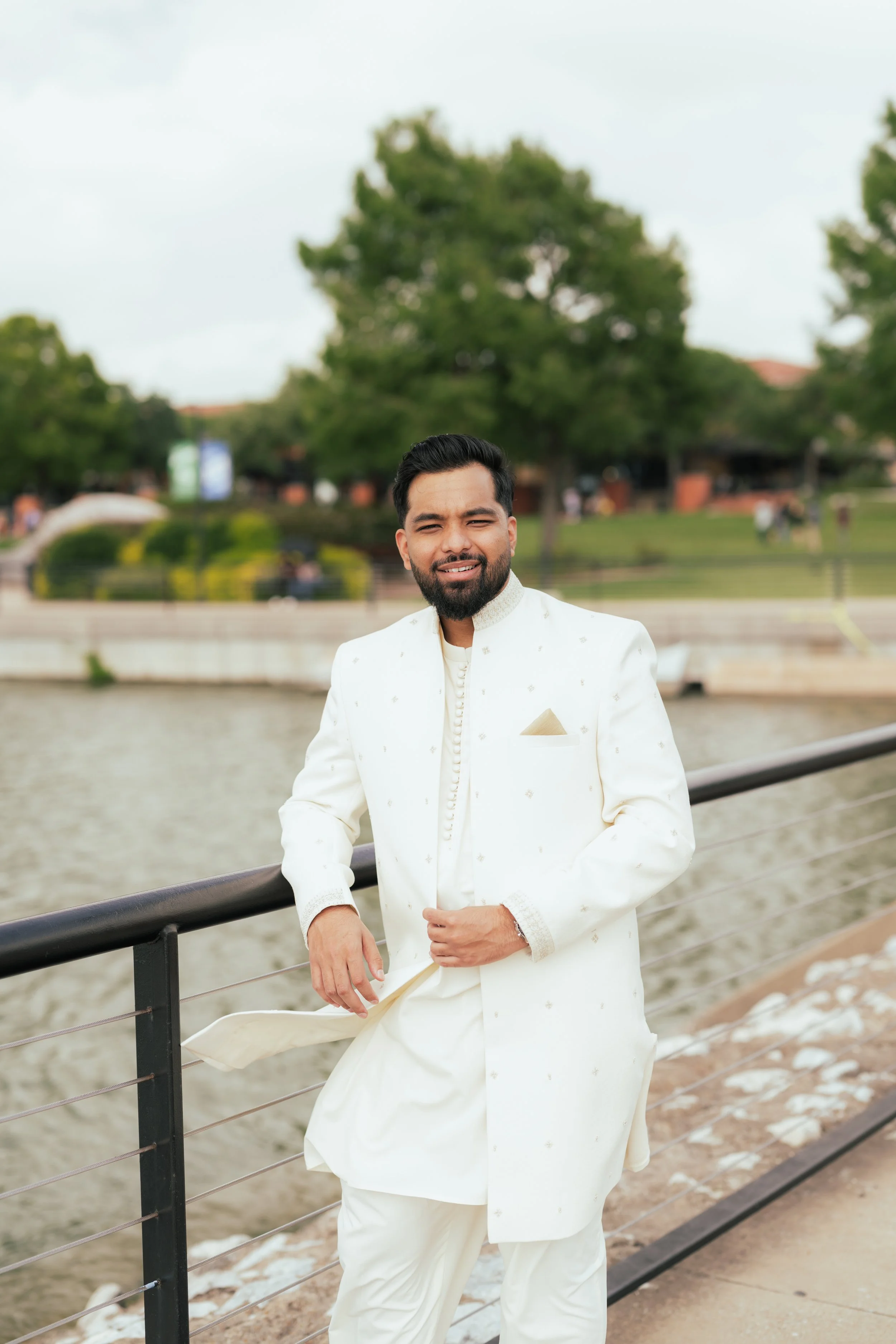 Man dressed in traditional white attire standing near a river with trees in the background.
