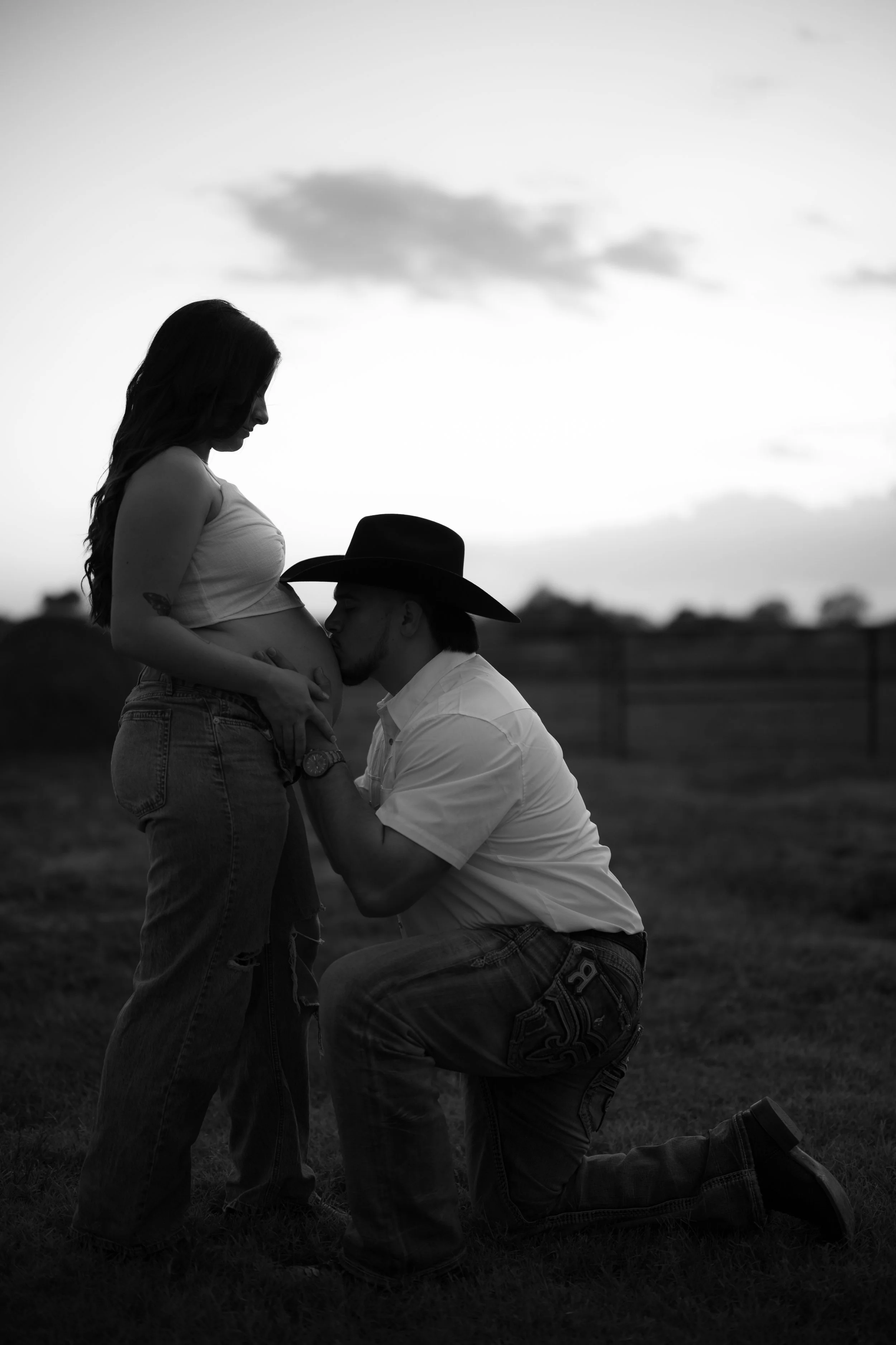 A black-and-white photo of a man kneeling and kissing a pregnant woman’s belly outdoors during sunset, with a cloudy sky in the background.