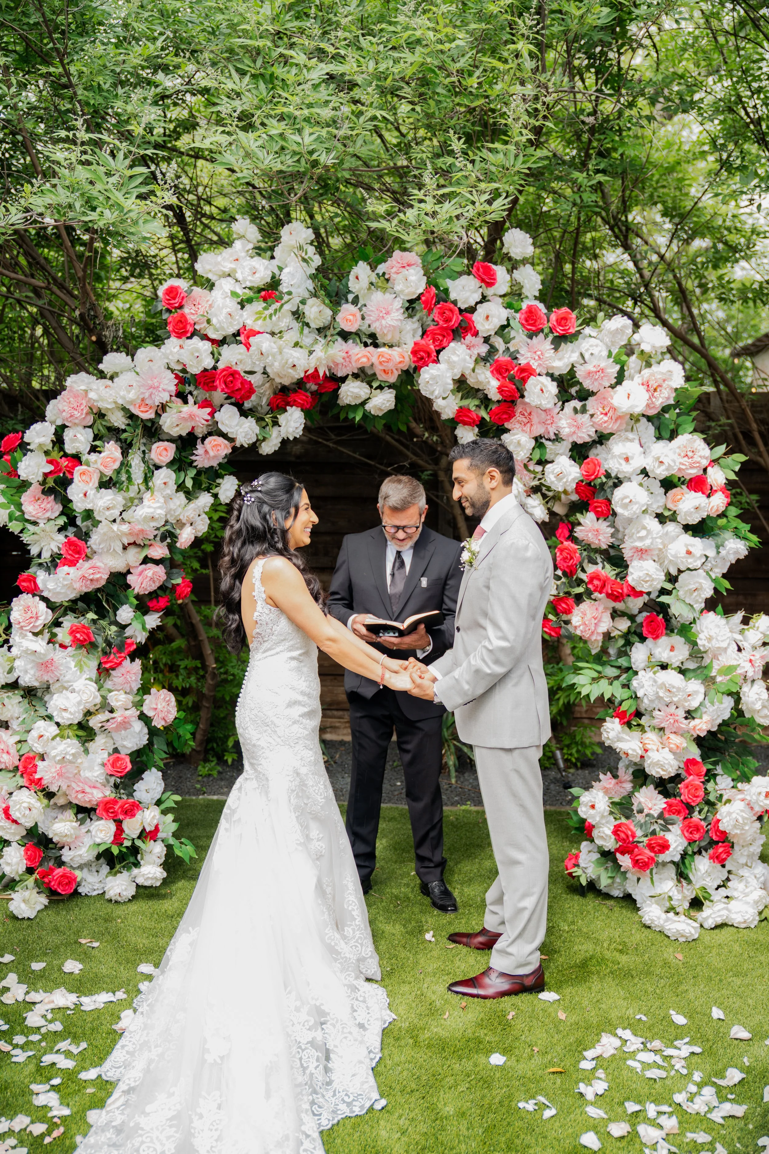 A bride and groom holding hands during their outdoor wedding ceremony, standing under a floral arch with pink and white flowers, in front of a green leafy background, with an officiant officiating.