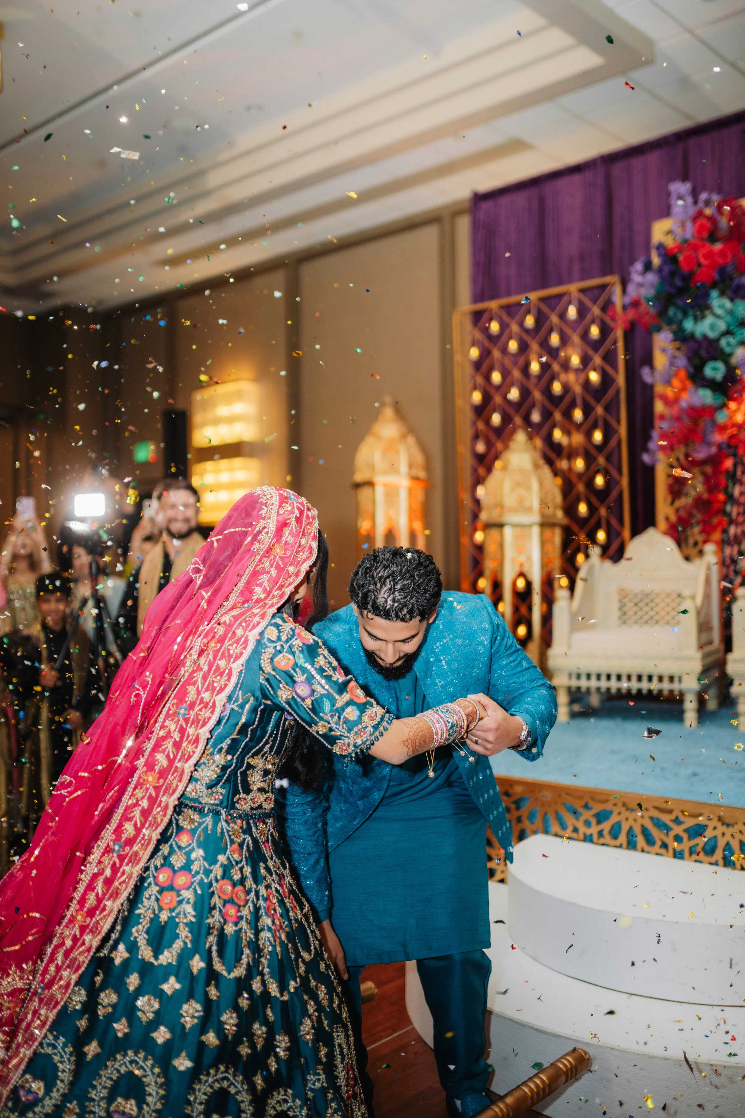 A couple in traditional Indian attire engaged in a wedding ceremony, with confetti falling around them and guests in the background.