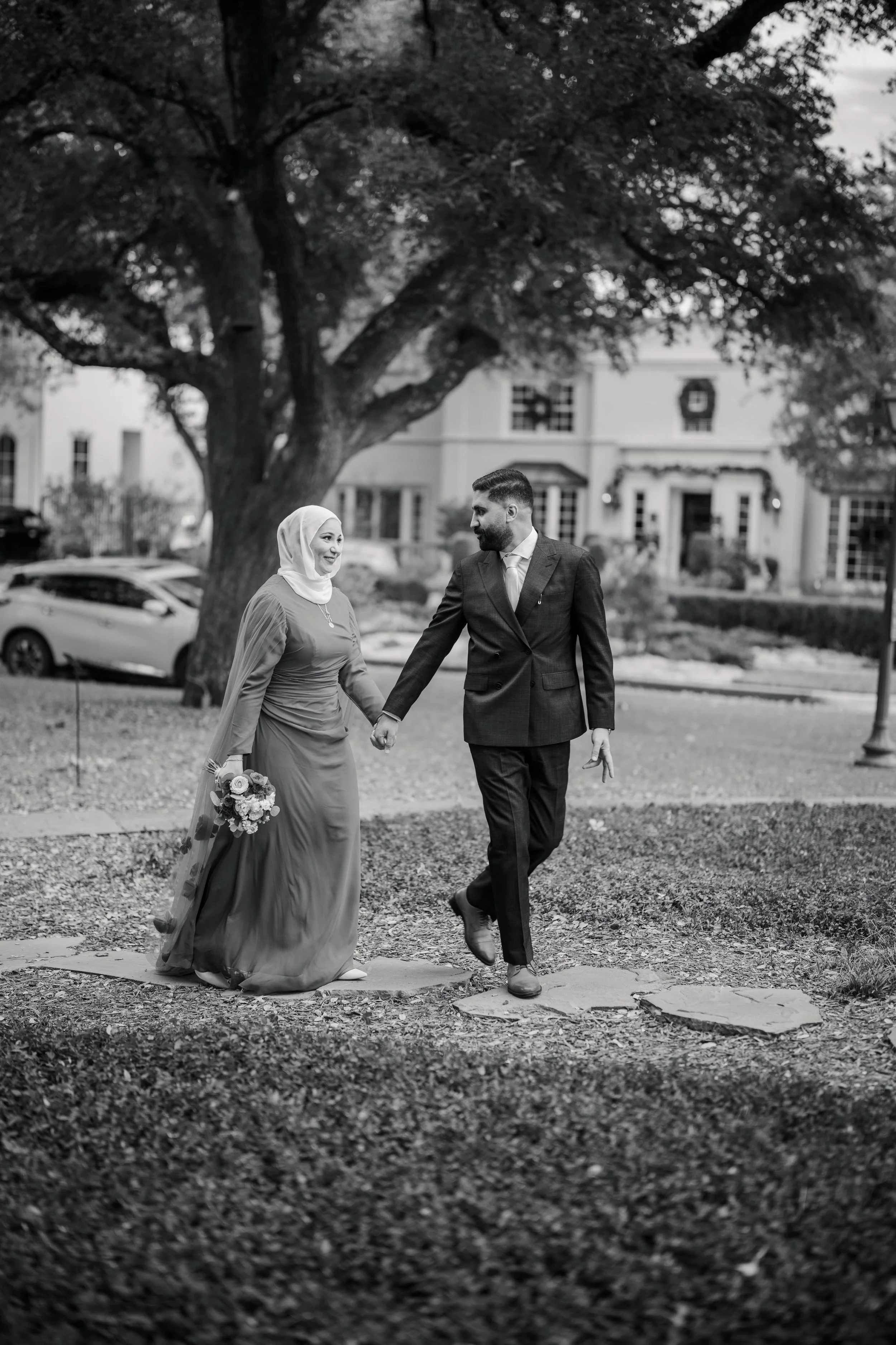 A black and white photo of a couple walking hand in hand outdoors, with the woman in a long dress and hijab holding a bouquet, and the man in a suit, under a large tree in a residential neighborhood.