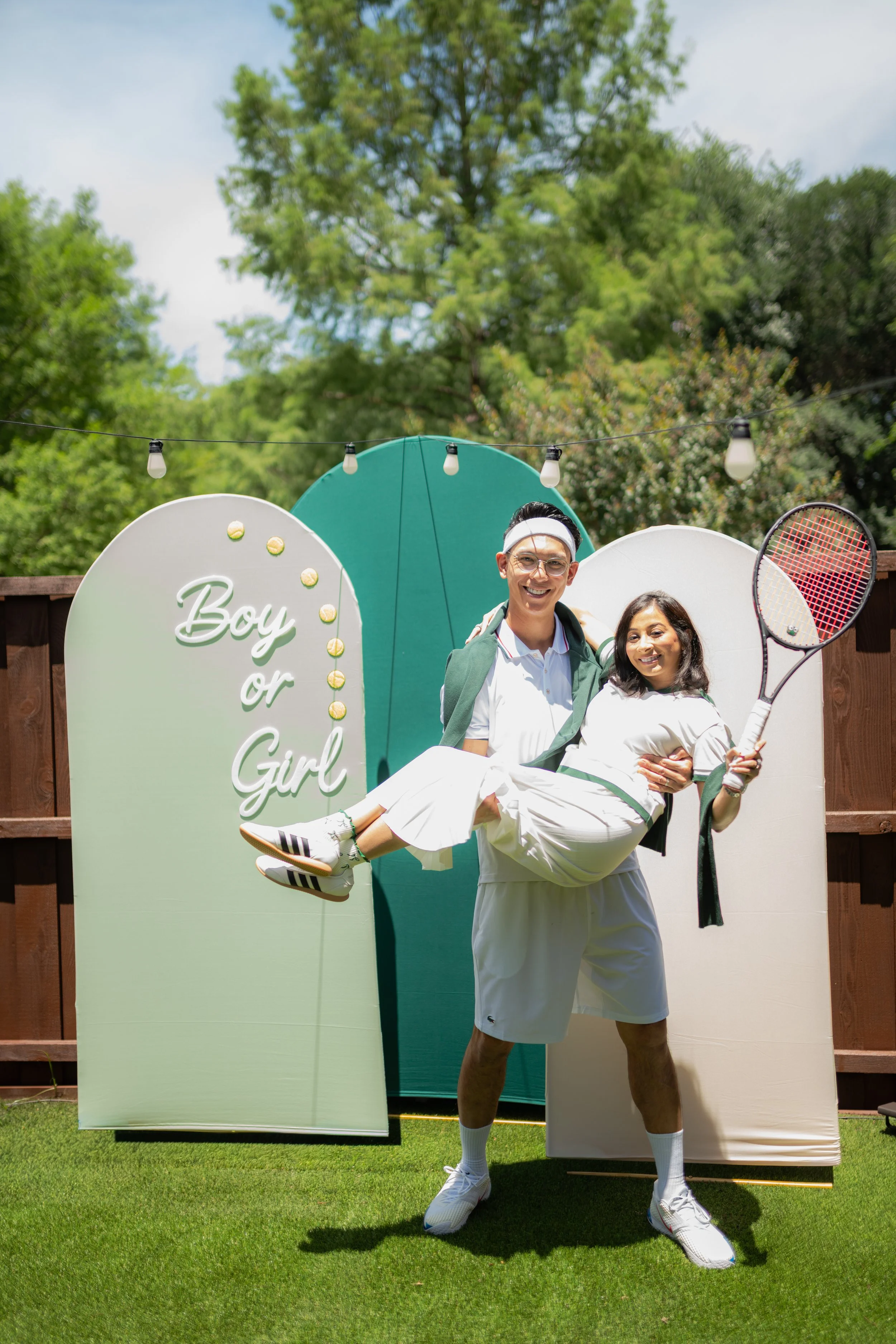 Two men, one holding a tennis racket and the other being carried, standing in front of a backdrop with signs that say "Boy or Girl" and tennis balls, outdoors on a lawn with trees in the background.