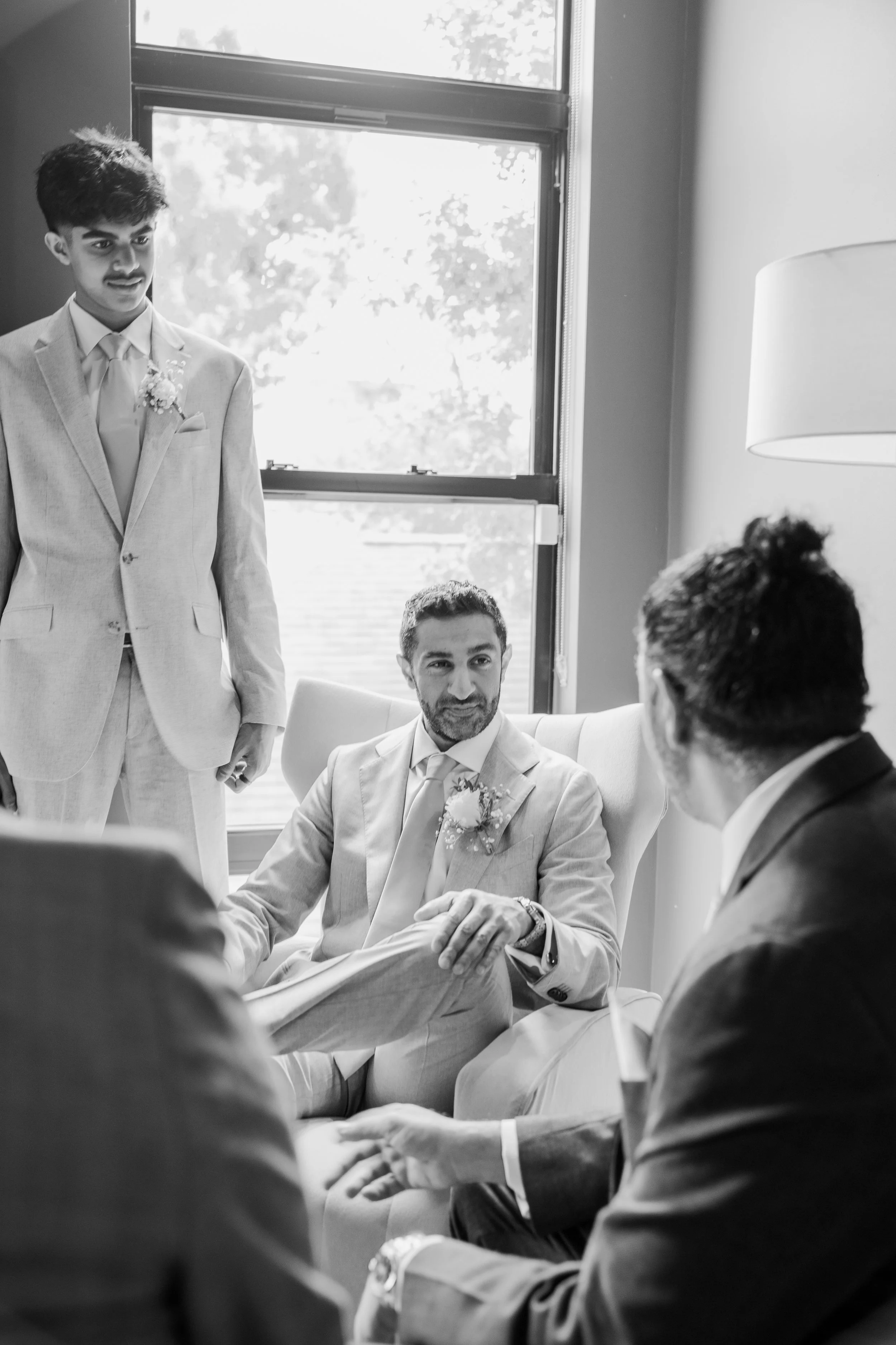 A black-and-white photo of a groom in a suit with boutonniere, sitting at a table during a wedding reception, with as many as six men around him engaged in conversation, in a room with a large window and natural daylight.
