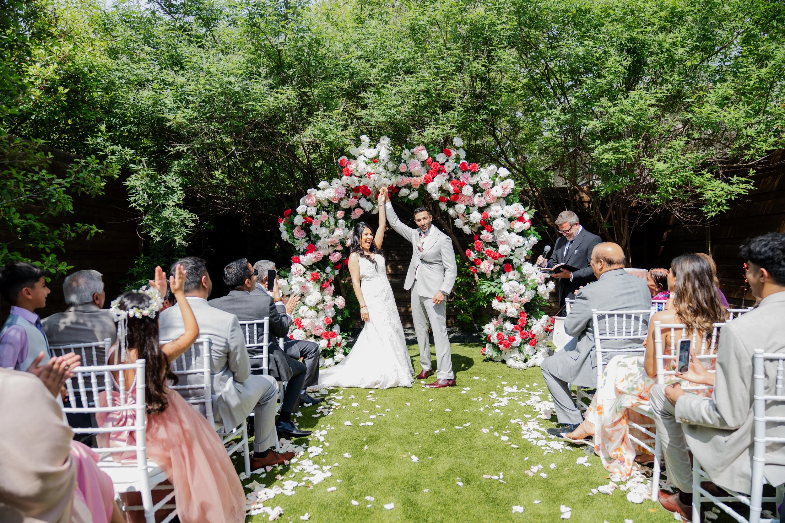 A wedding ceremony taking place outdoors with the bride and groom holding hands under a floral arch, surrounded by seated guests, on a sunny day with green trees in the background.