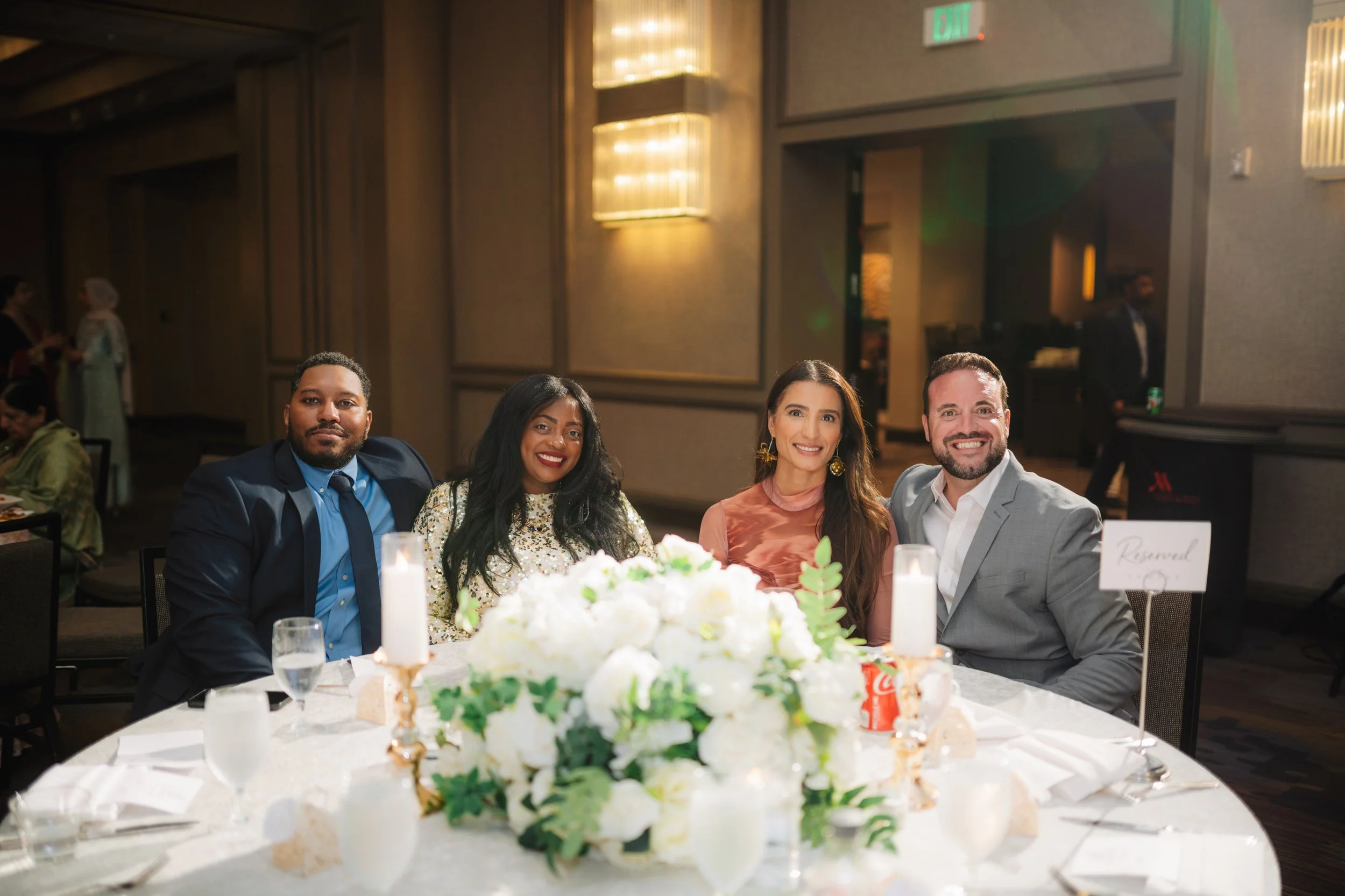 People sitting at a round table during a celebration or event, with floral centerpiece and candles, in a banquet hall setting.