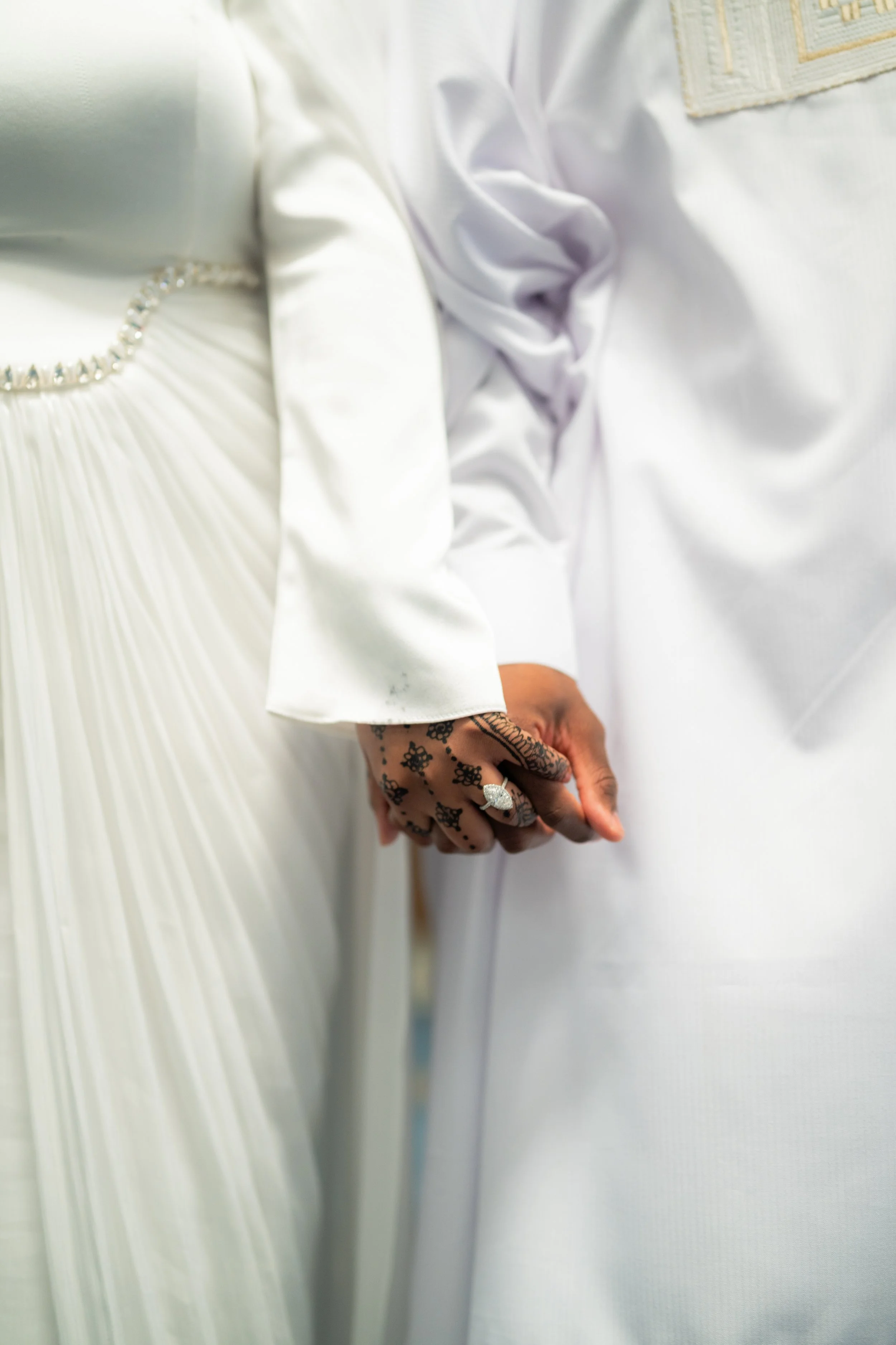 Close-up of two people holding hands, both dressed in white, with one person wearing a wedding ring and intricate henna tattoos.