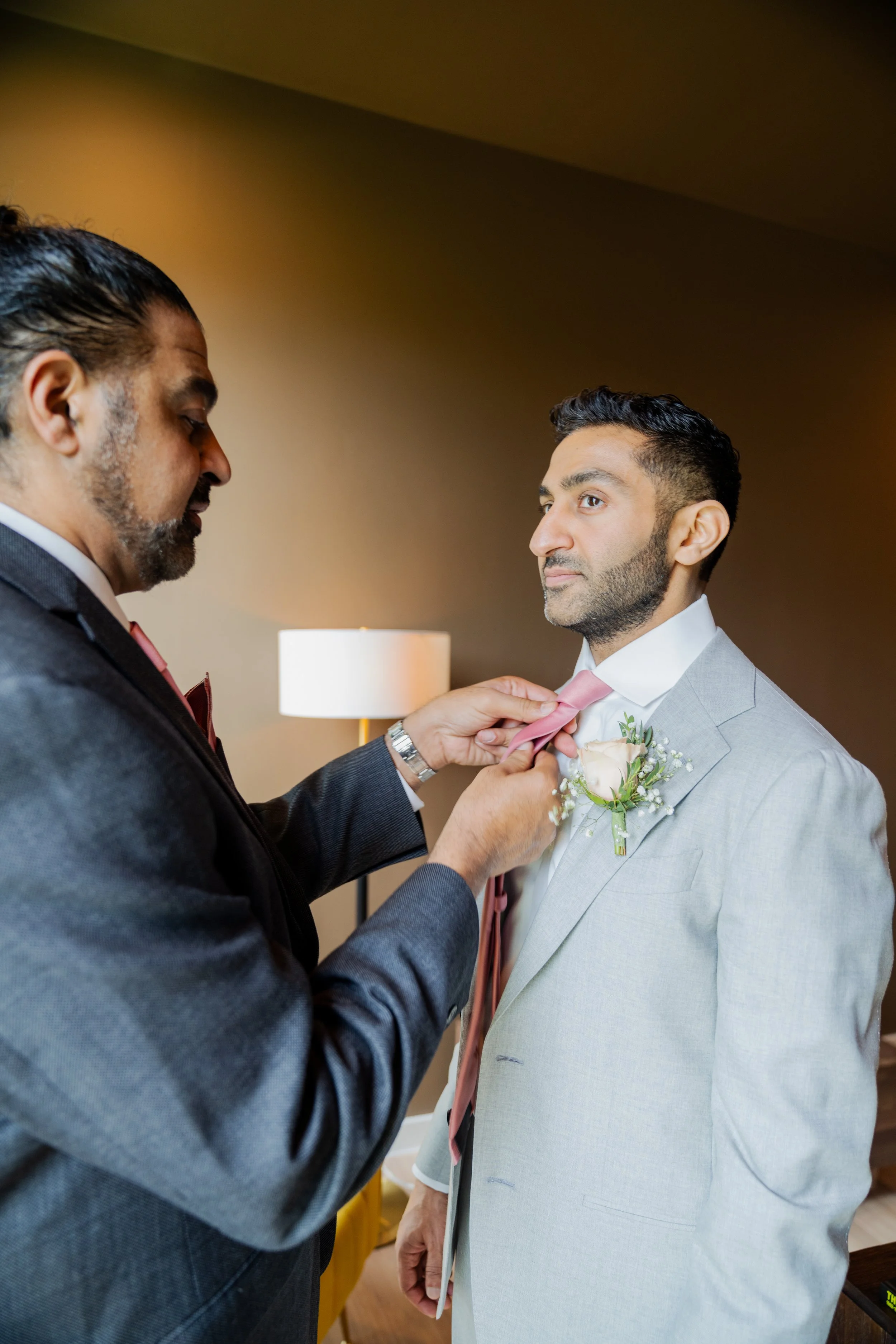 A man in a light gray suit with a pink tie is being assisted by another man in a dark suit with a pink tie, who is adjusting his tie during a wedding ceremony.