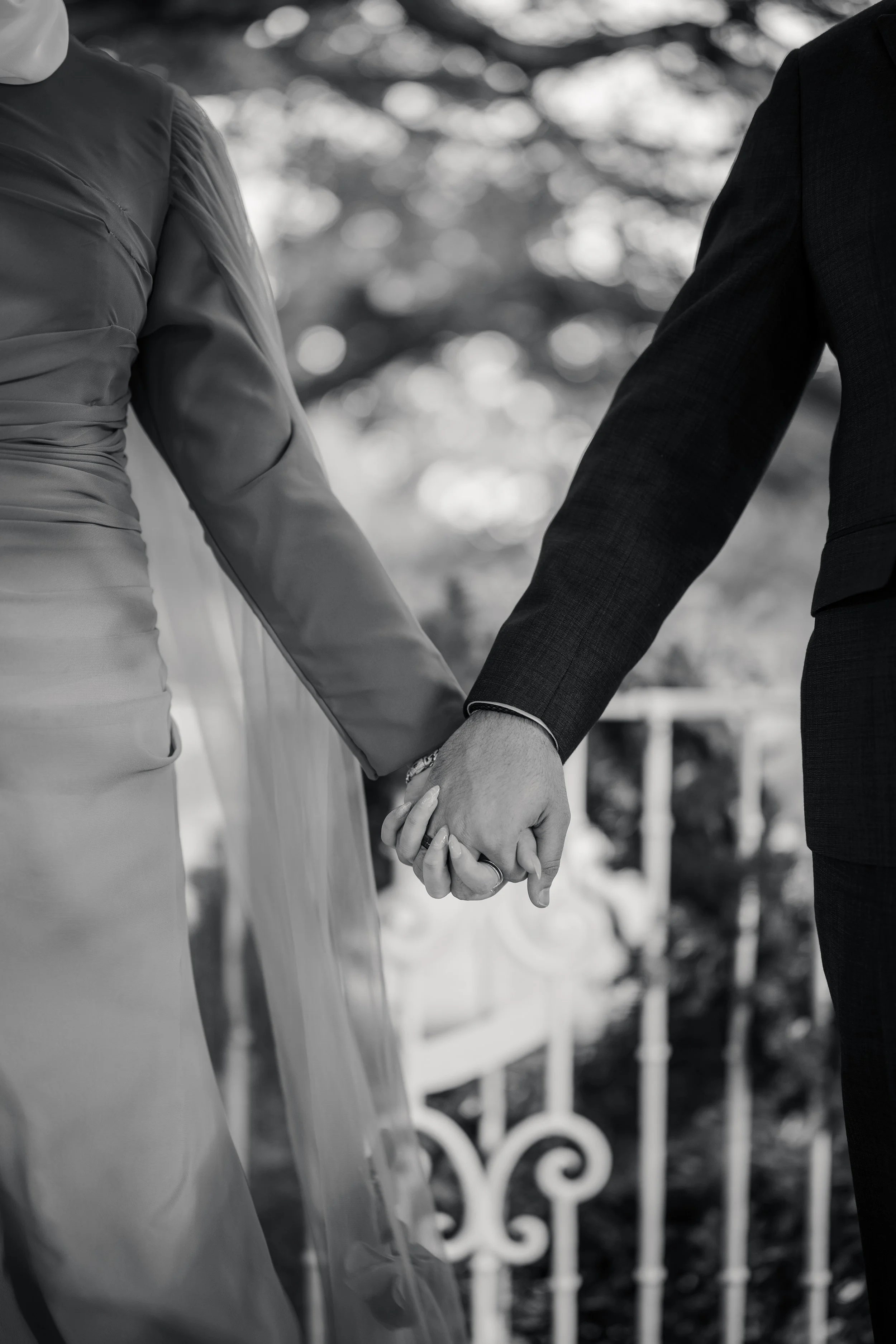Close-up of a bride and groom holding hands during a wedding ceremony. The bride wears a satin dress with long sleeves, and the groom wears a dark suit. The background is blurred with trees and decorative fencing.