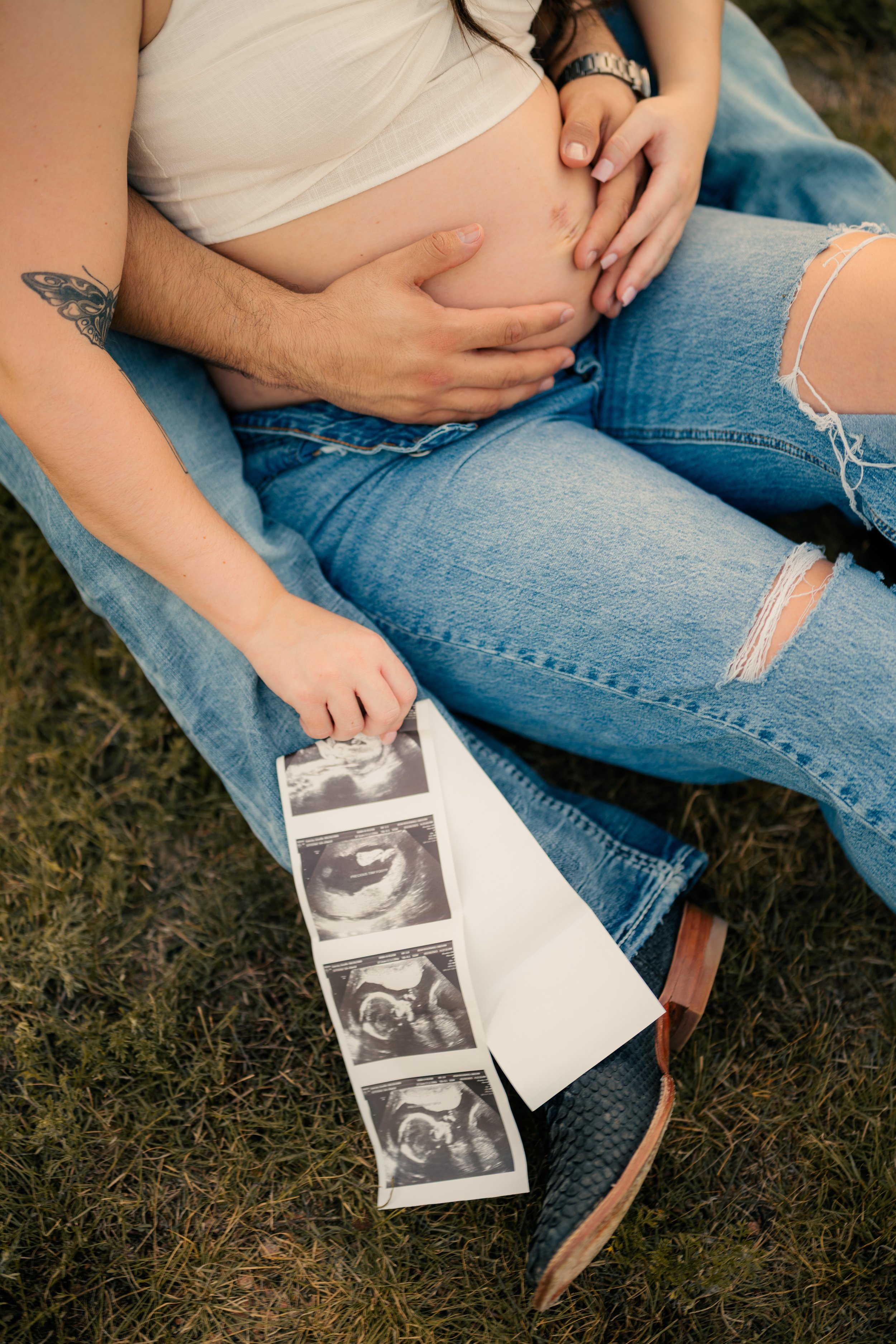 A pregnant woman holding ultrasound pictures while sitting on grass, with a partner's hand on her belly.
