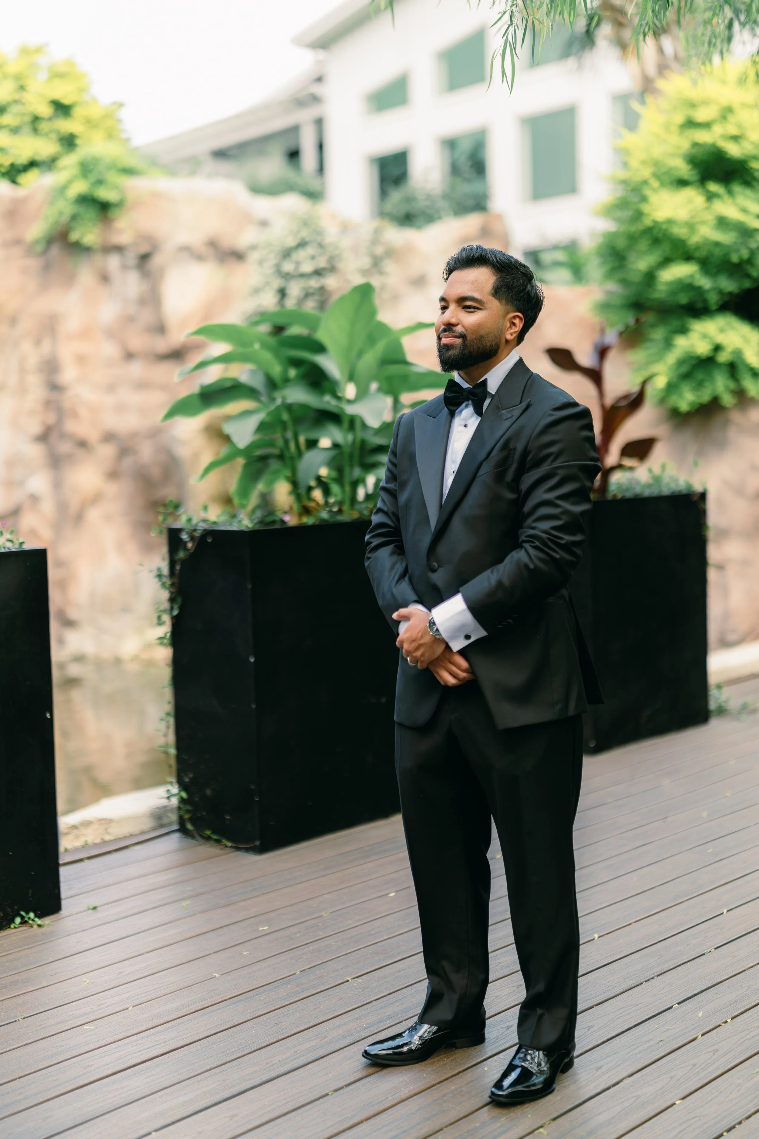 Man dressed in a black tuxedo with a bow tie standing outdoors on wooden decking, surrounded by large potted plants and greenery, with a modern house in the background.
