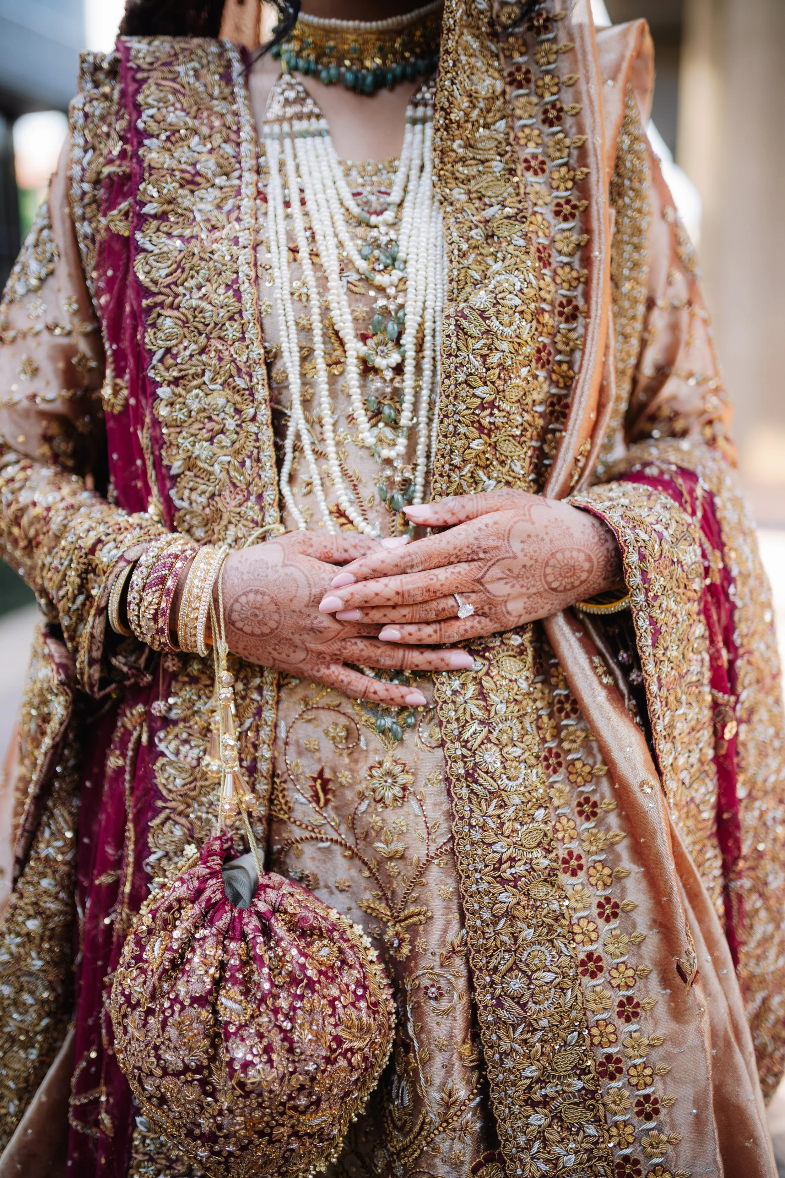 Close-up of a woman wearing ornate traditional Indian attire, including a heavily embroidered dupatta with gold floral and leaf patterns, layered pearl necklaces with floral accents, bangles, and henna designs on her hands.
