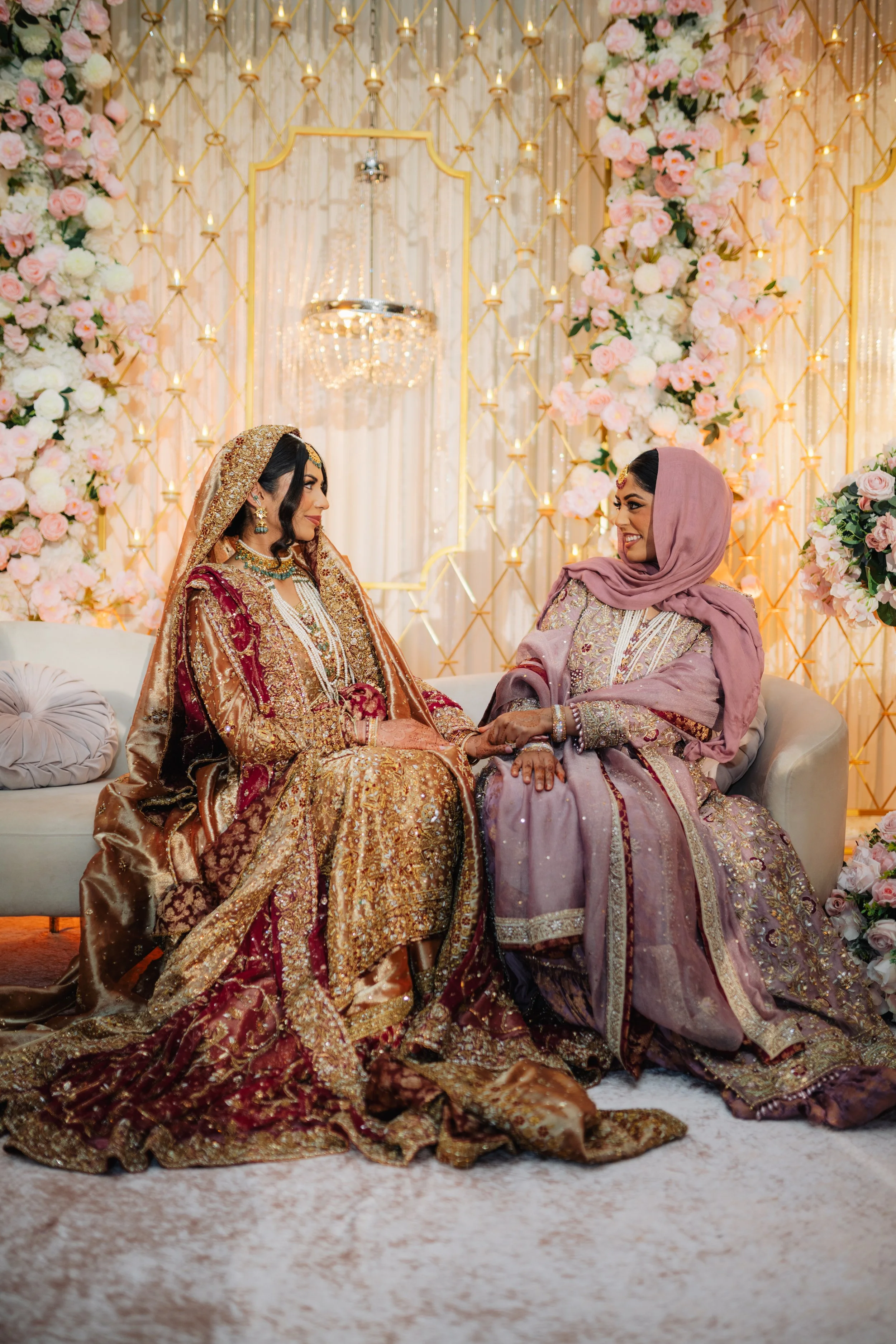 Two women dressed in traditional South Asian wedding attire, sitting on a sofa and holding hands, with ornate floral and gold decor in the background.