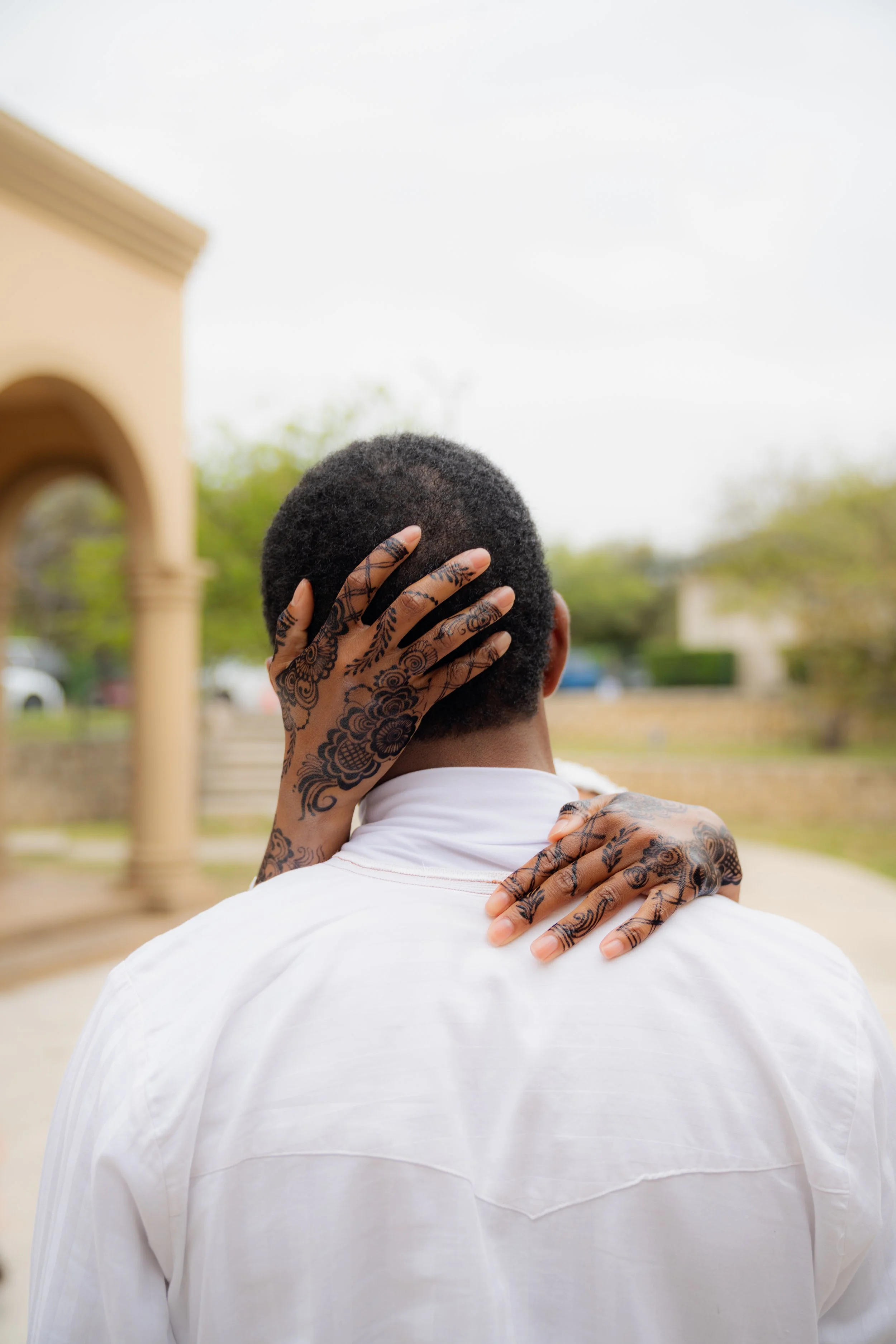 Person with tattoos on hand and neck, seen from behind, holding their head and shoulder outdoors during daytime.