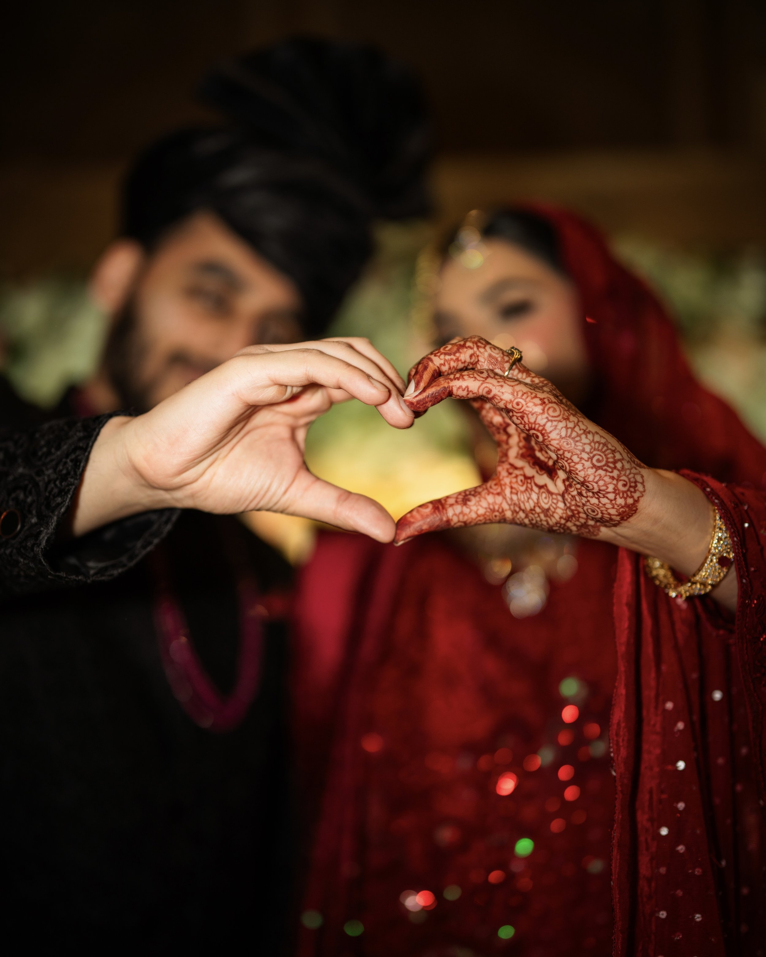 A couple forming a heart shape with their hands during a wedding celebration. The woman wears traditional Indian attire and mehndi (henna) on her hand, and the man is dressed in traditional Indian wedding clothing.