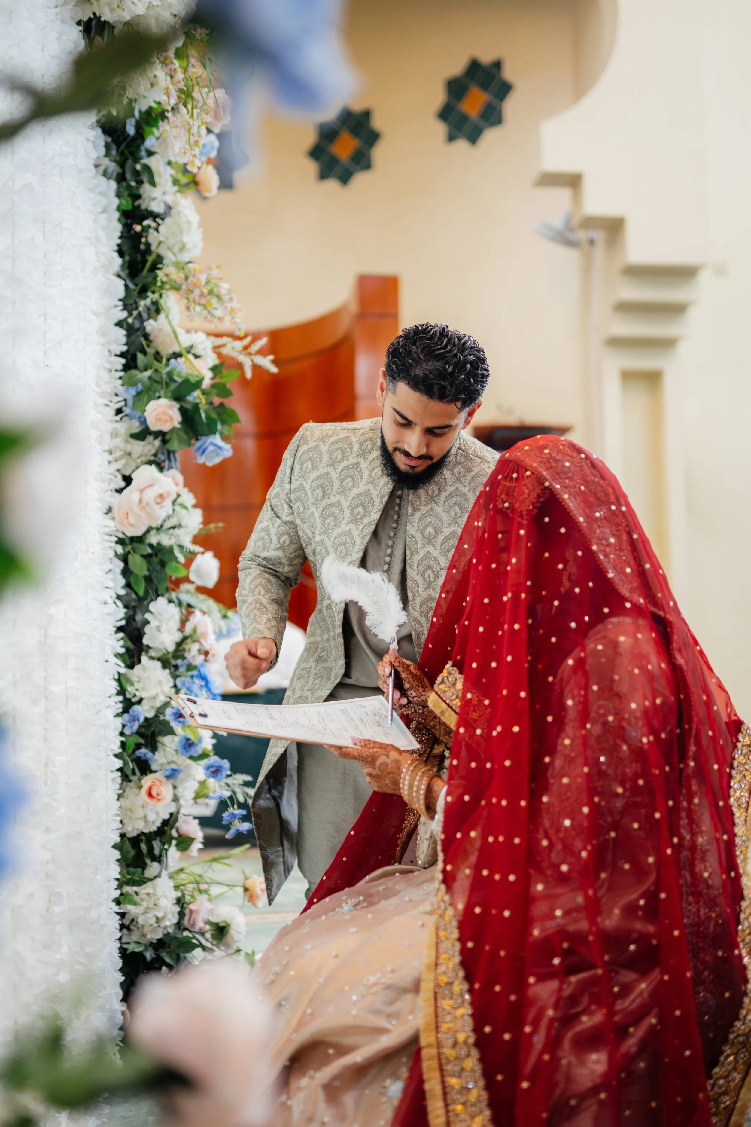 A bride and groom during their wedding ceremony, with the bride dressed in a traditional red and gold outfit and the groom in a beige patterned jacket, standing in front of white and pastel floral arrangements.