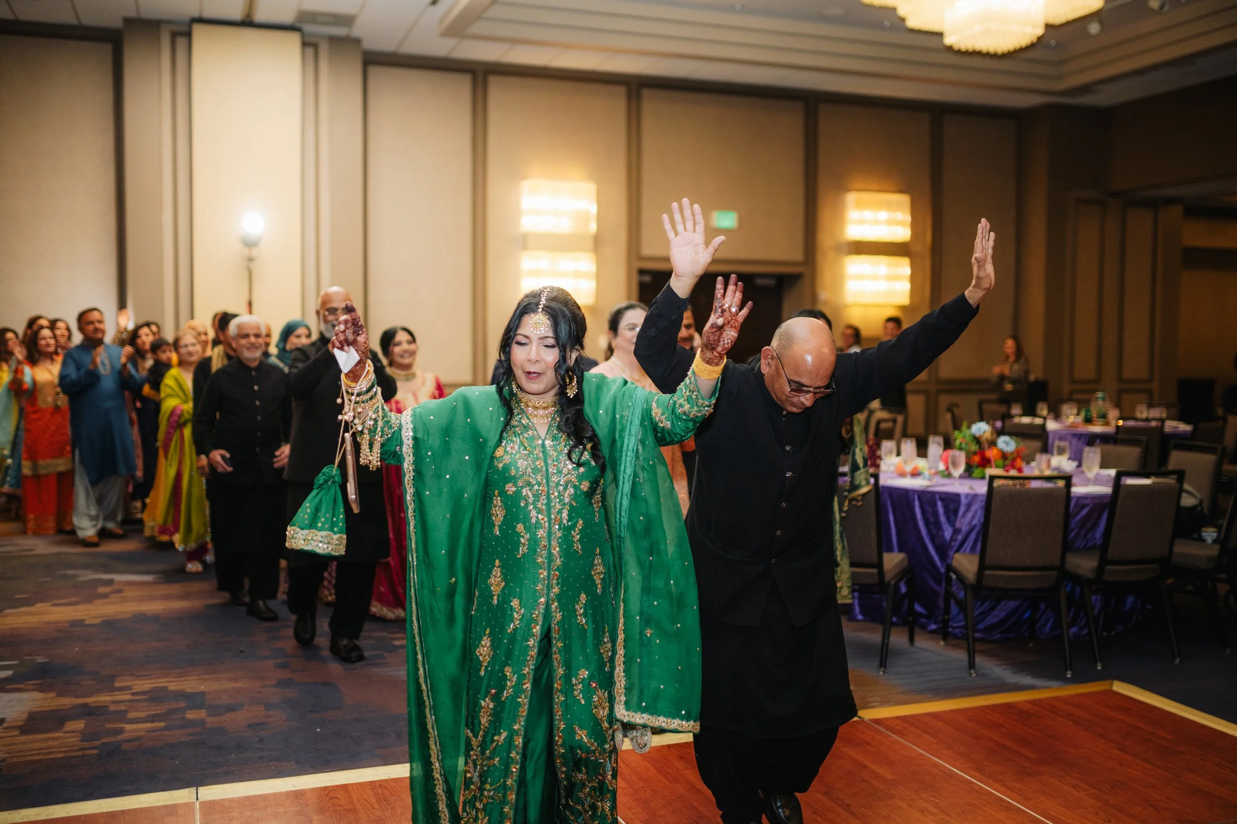 People participating in a dance or celebration at a formal event, with a woman in traditional green attire and a man in black in the foreground.