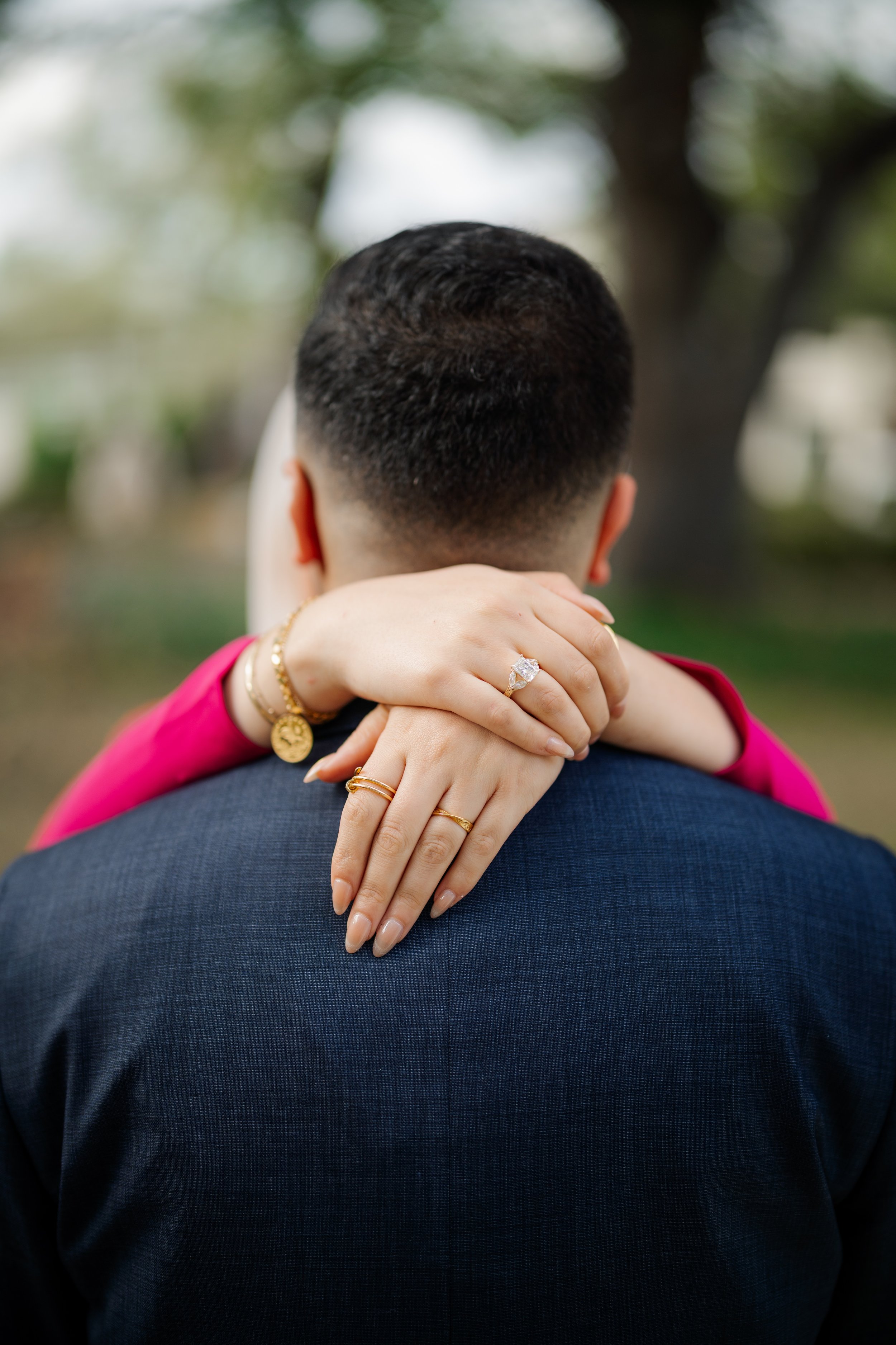 A woman with light skin embracing a man with light skin from behind, her arms around his shoulders. She is wearing gold jewelry, including a ring with a large gemstone and a gold bracelet. The man is wearing a dark blue suit, and his face is not visi