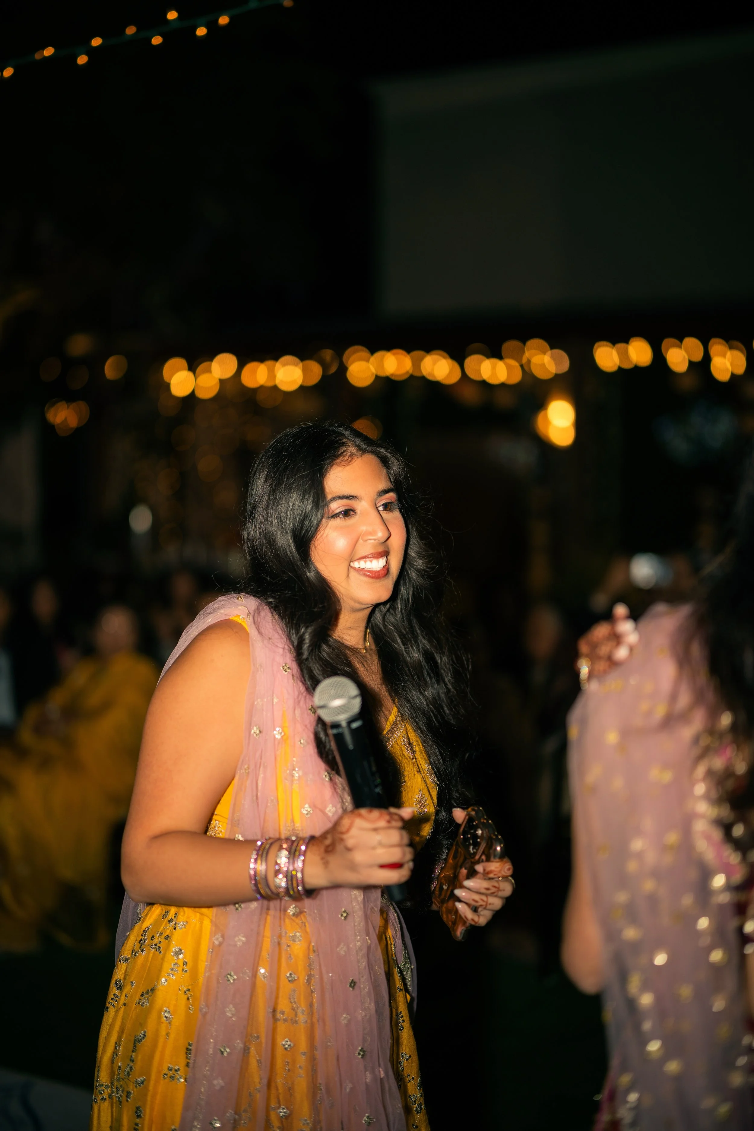 A woman with long black hair, wearing a yellow traditional Indian dress with pink and gold accents, holding a microphone and smiling at an outdoor evening event with fairy lights in the background.