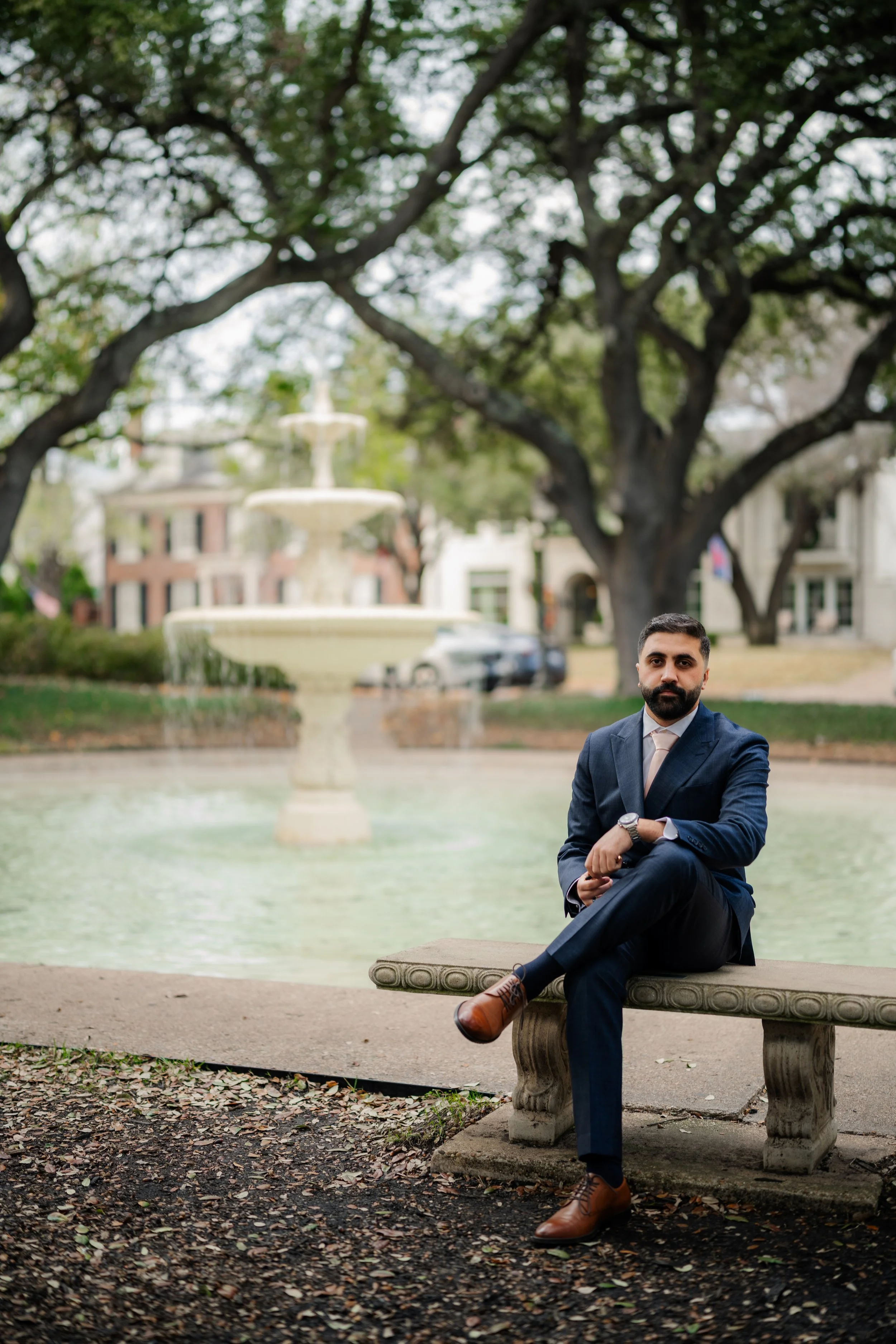 A man in a navy blue suit and brown shoes sitting on a stone bench in front of a fountain in a park, with large trees and buildings in the background.