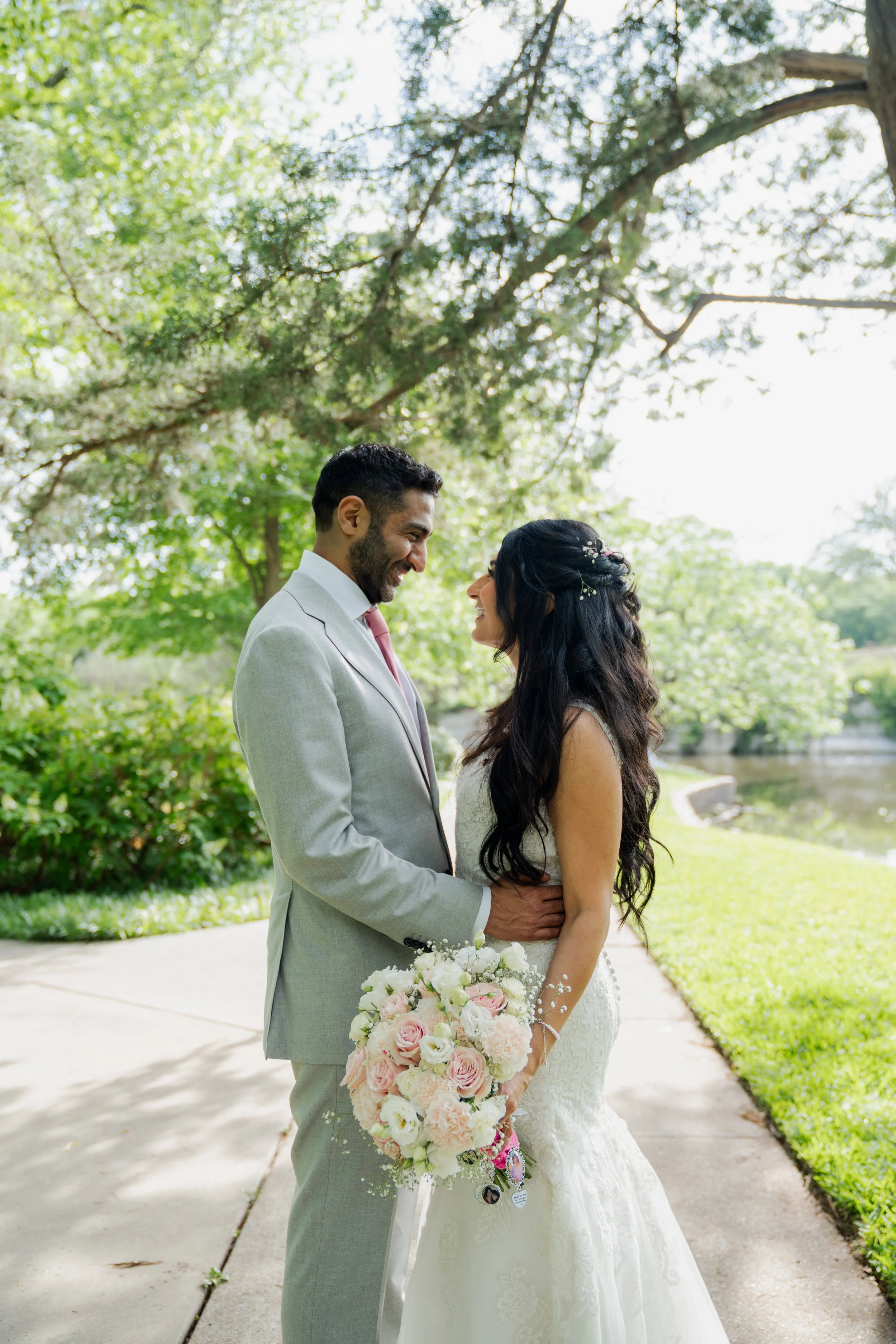 A bride and groom standing face to face outdoors on a cloudy day, smiling and holding hands. The bride has long dark hair and is wearing a white lace wedding dress, holding a bouquet. The groom has short dark hair and a beard, wearing a light gray su