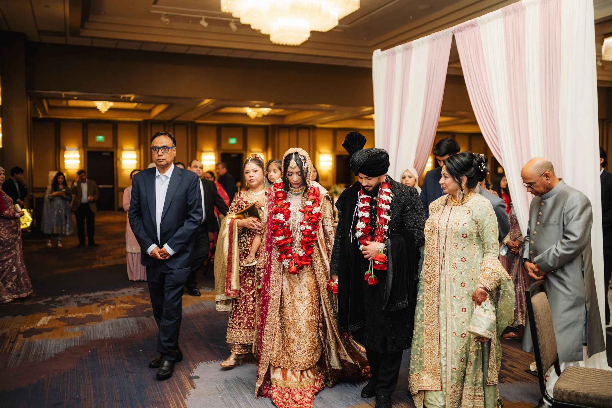 A group of people dressed in traditional South Asian attire participating in a wedding ceremony, with a couple in the center amidst garlands, under a decorated arch in a banquet hall.