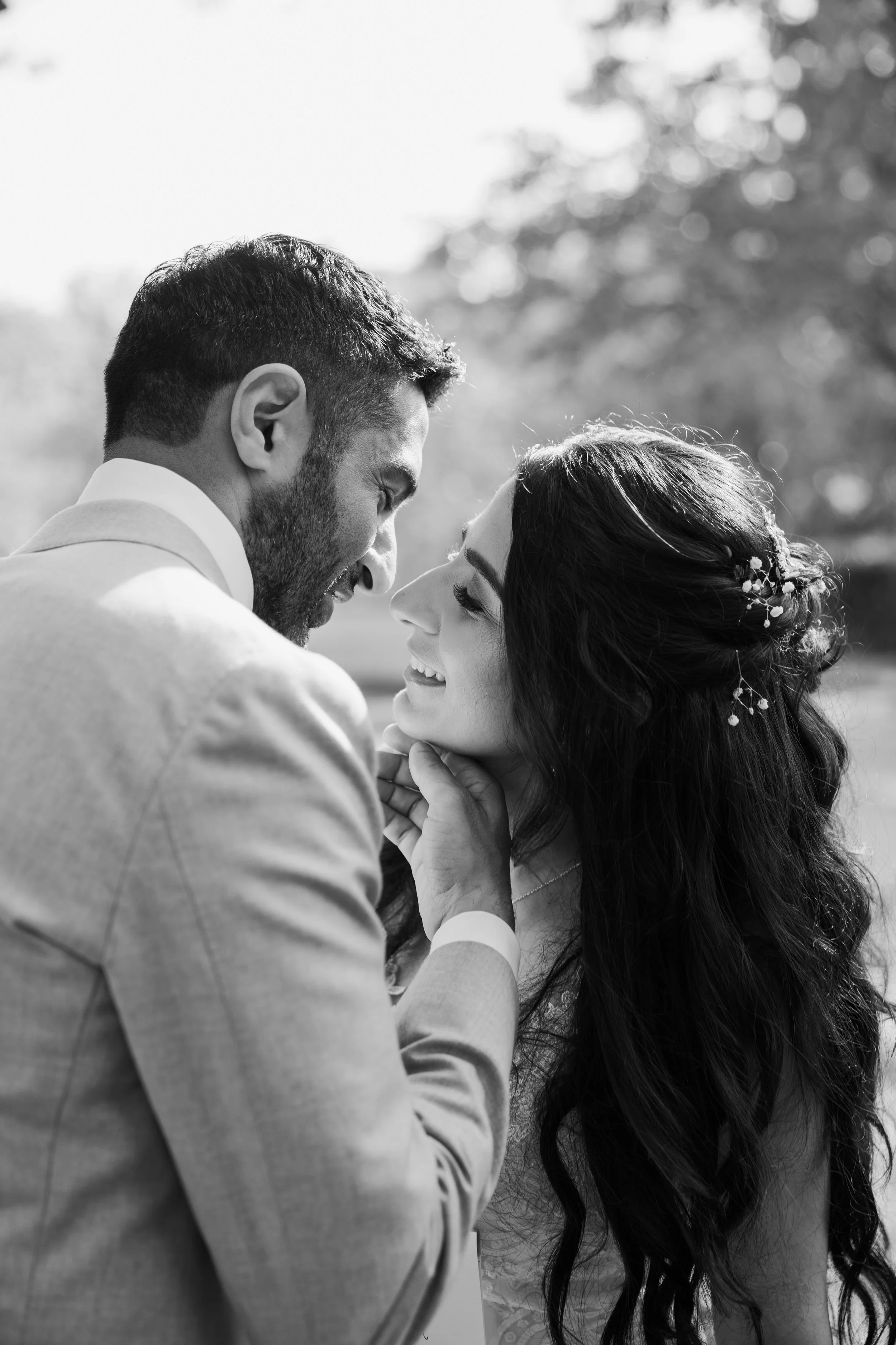 A black and white photograph of a couple smiling closely together outdoors, with blurred trees in the background. The man has short hair and a beard, and the woman has long, wavy hair with floral hair accessories.